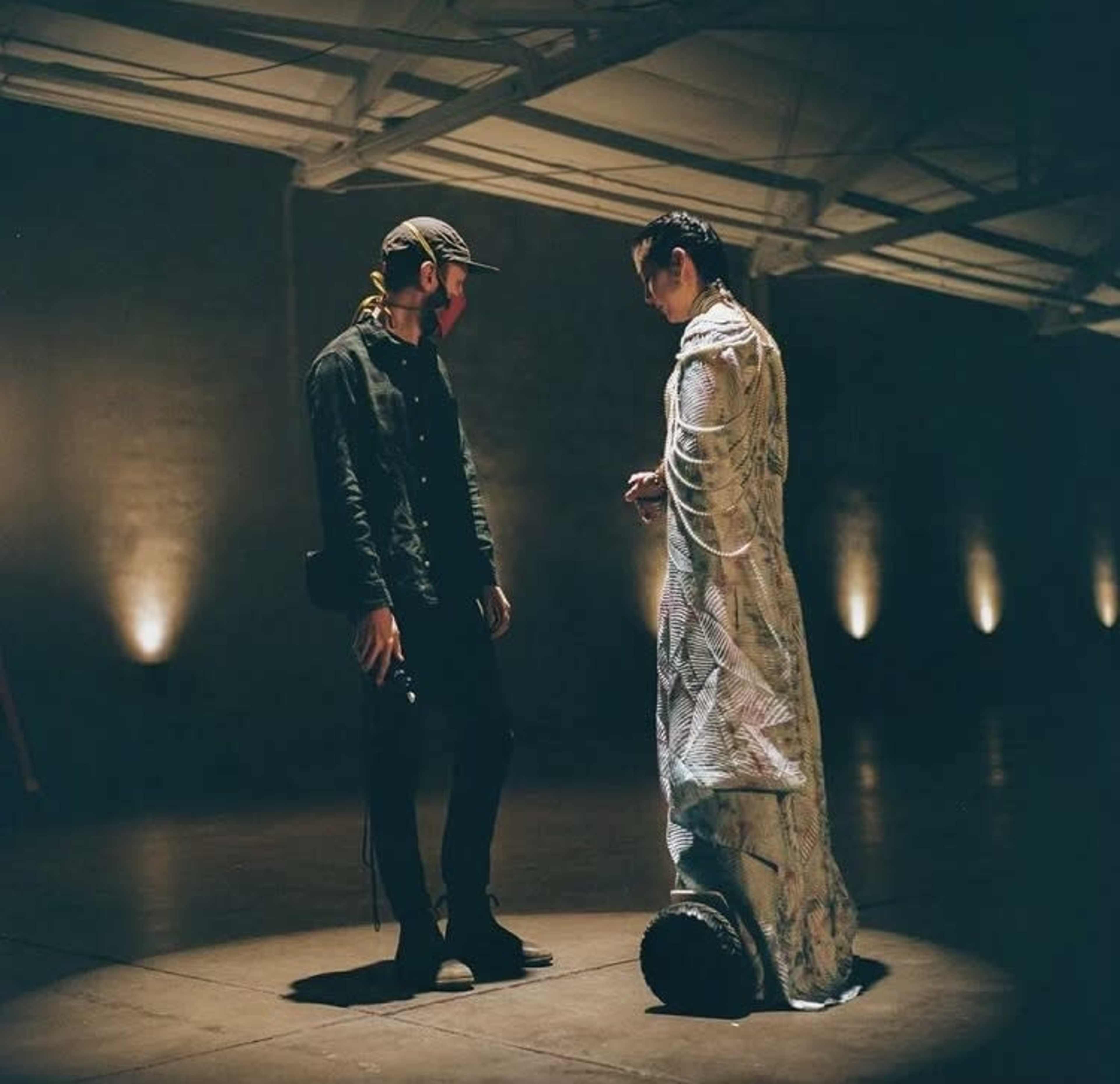 A man in dark clothing stands conversing with a person dressed in a decorative, flowing garment in an industrial space with subdued lighting.