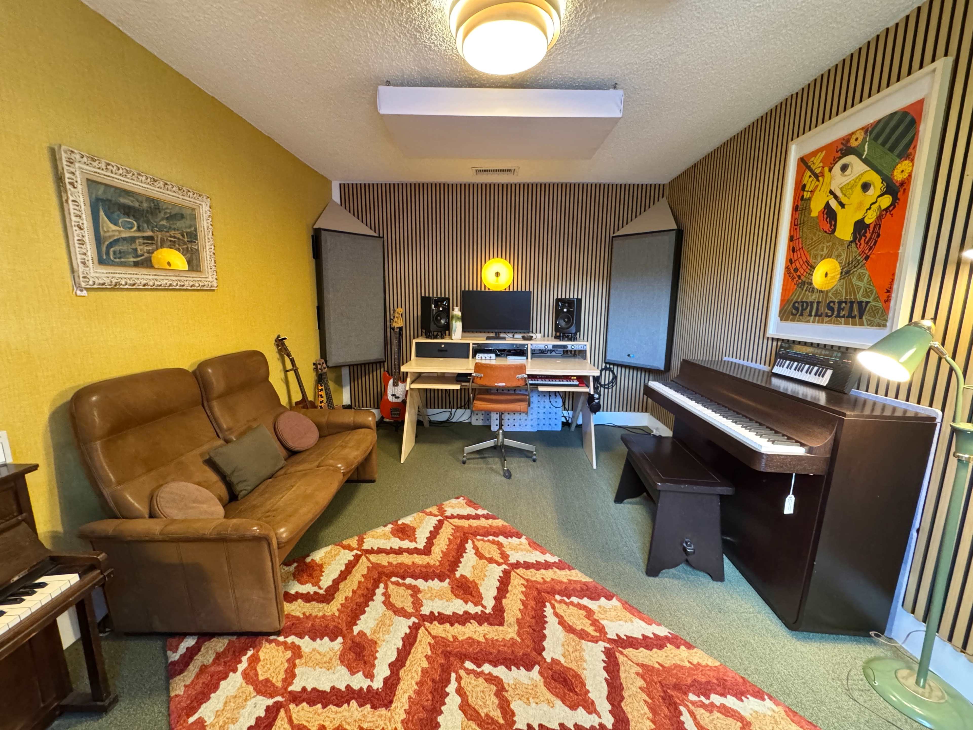 The image shows a cozy music studio featuring a brown sofa, a computer workstation with speakers, a piano, and a colorful rug against a yellow and striped wall.
