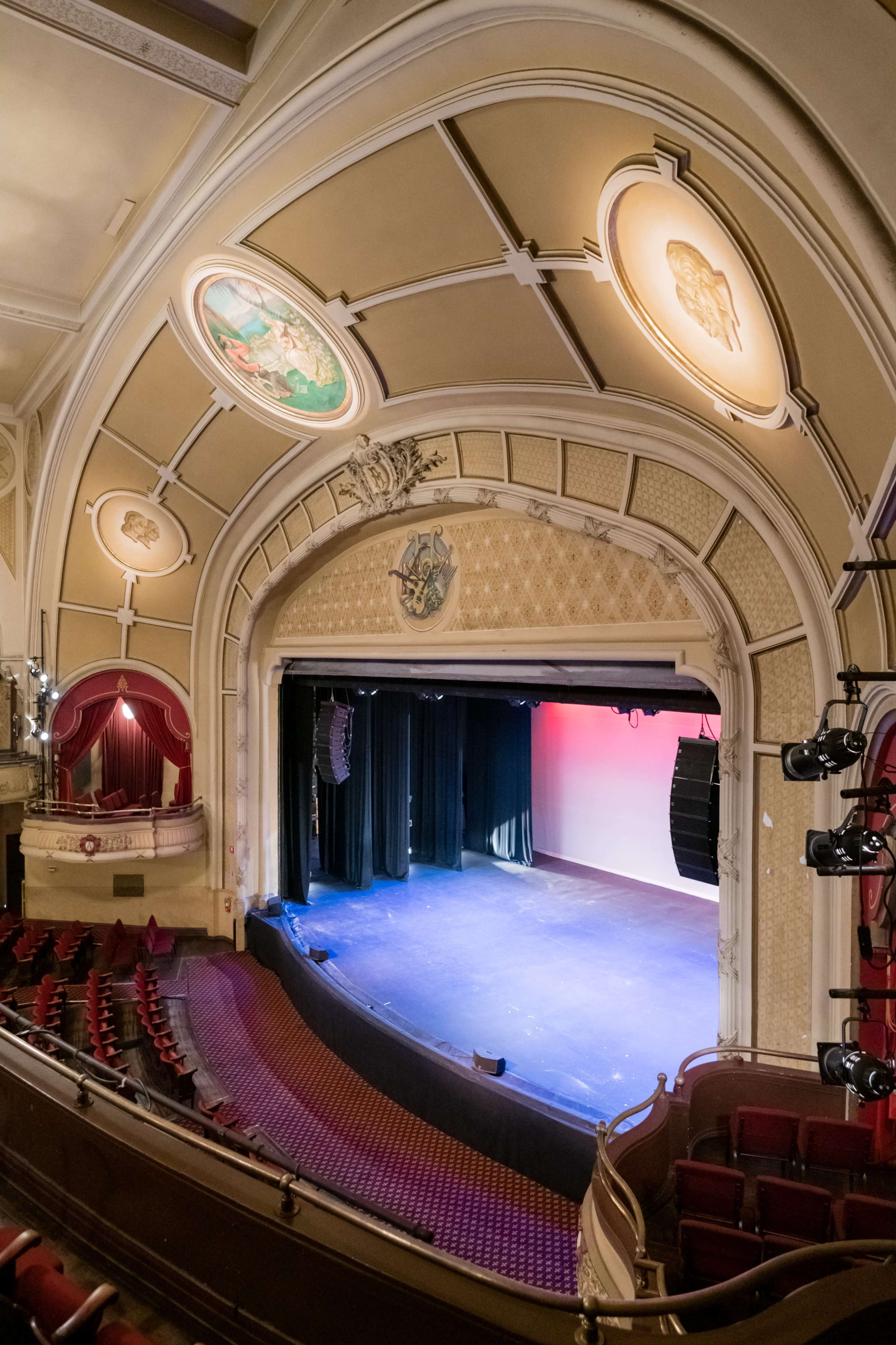 The image shows an ornate theater interior with a stage illuminated by colorful lights and elegant architectural details in the ceiling.
