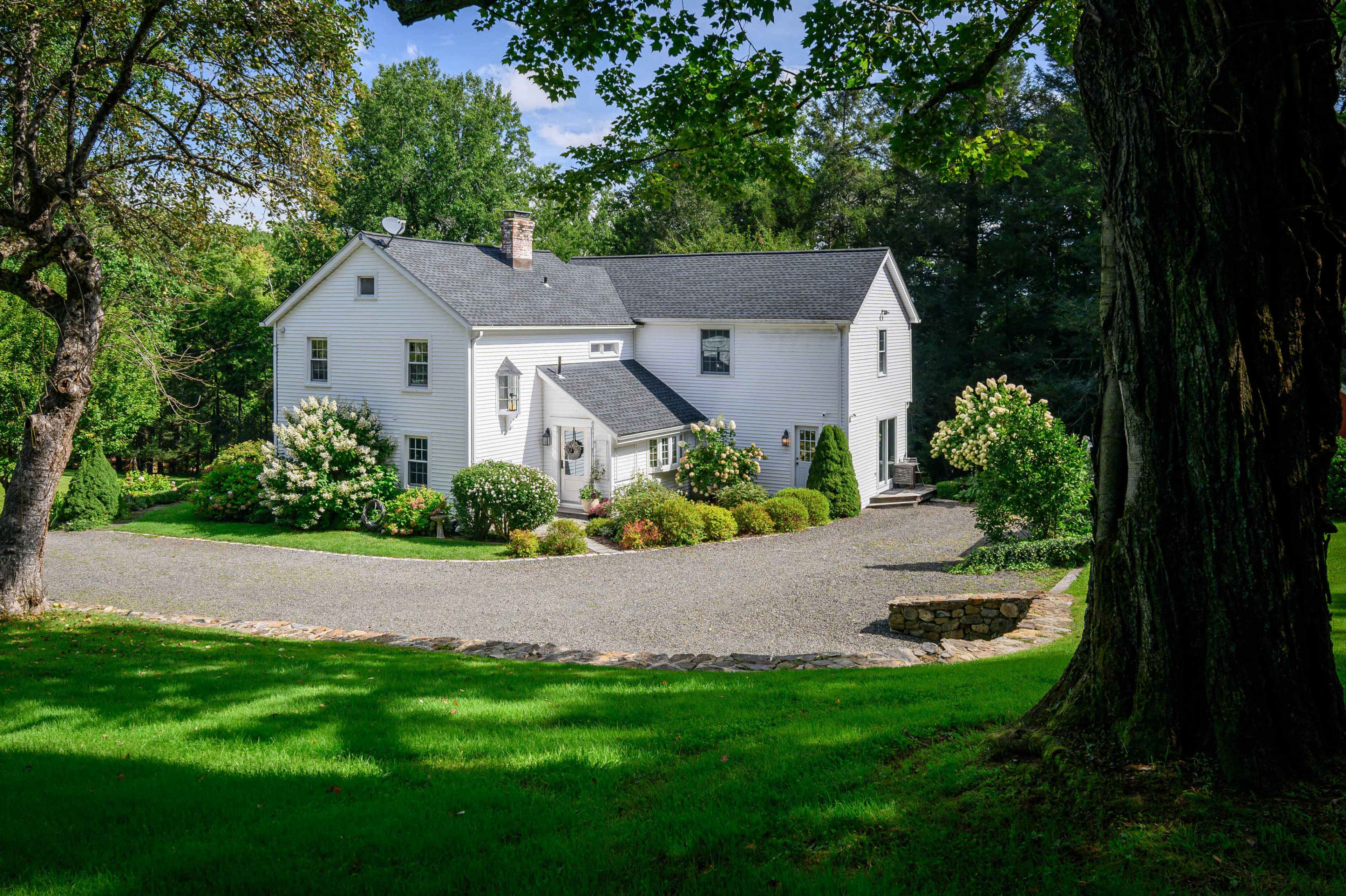 The image shows a white two-story house surrounded by green grass and lush trees, with a gravel driveway leading up to it.