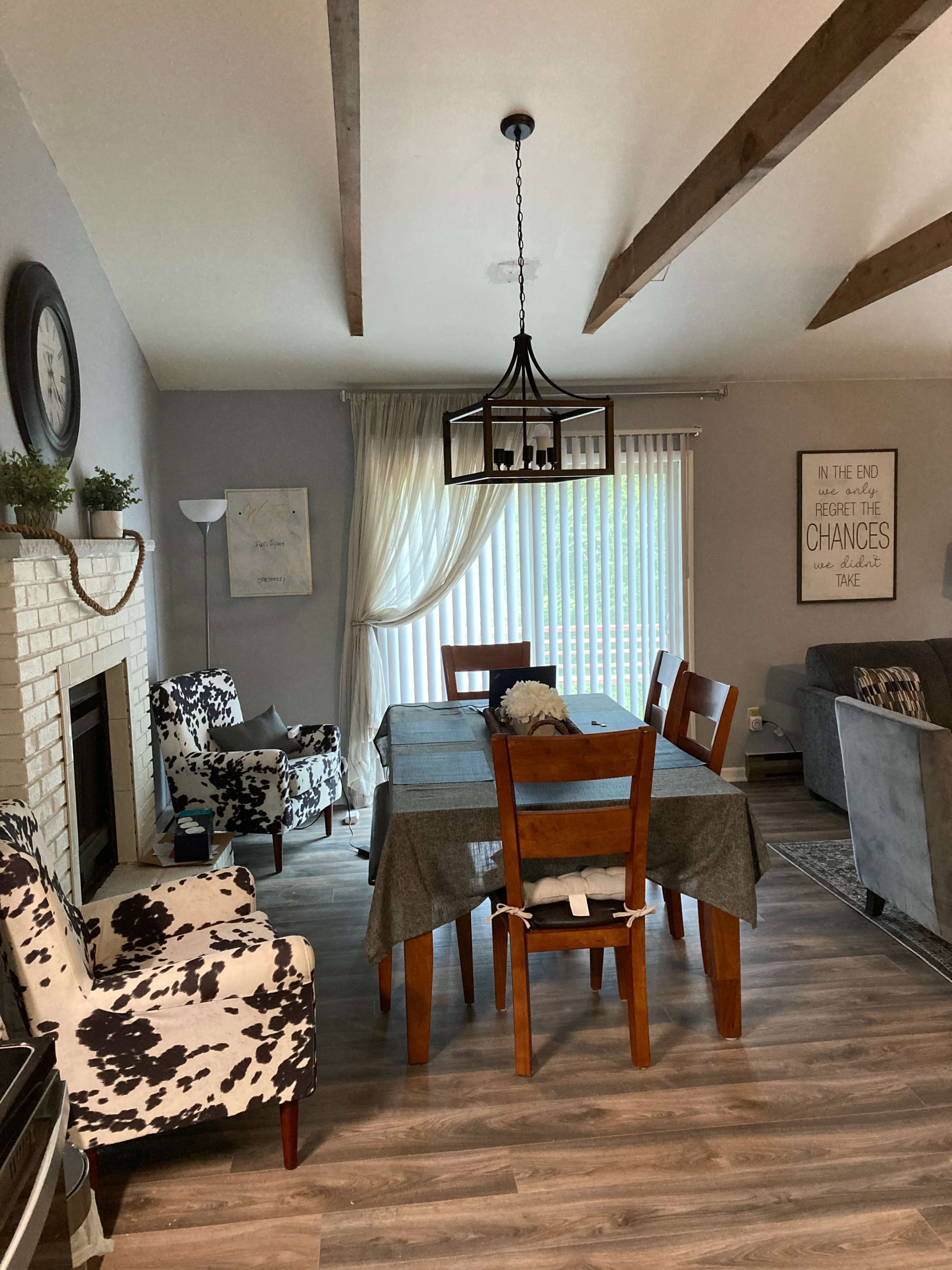 A dining area with a table surrounded by wooden chairs and patterned armchairs, featuring a fireplace and large window with sheer curtains.