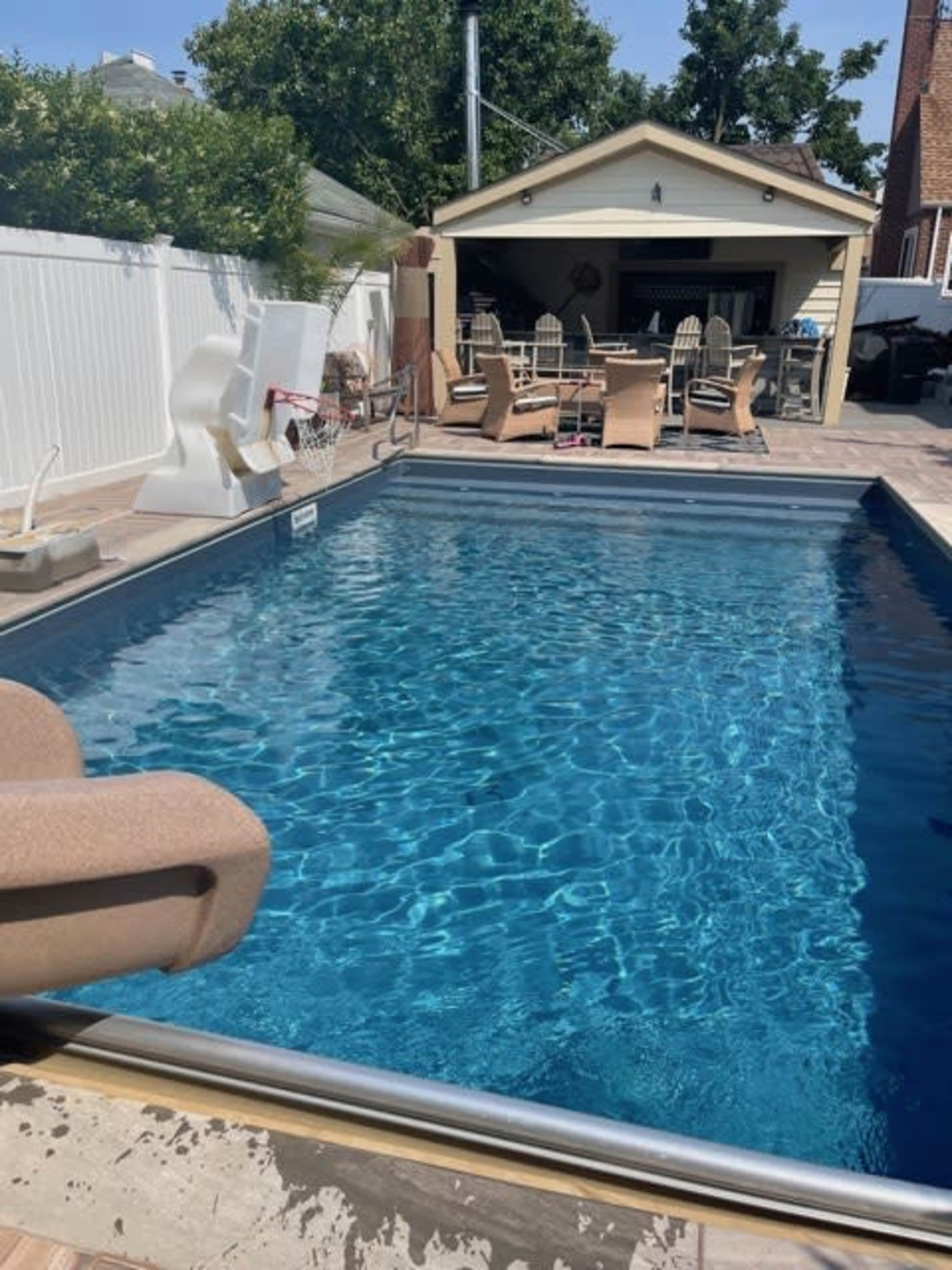 The image shows a clear swimming pool with a slide beside a patio area featuring outdoor furniture and a shaded structure.