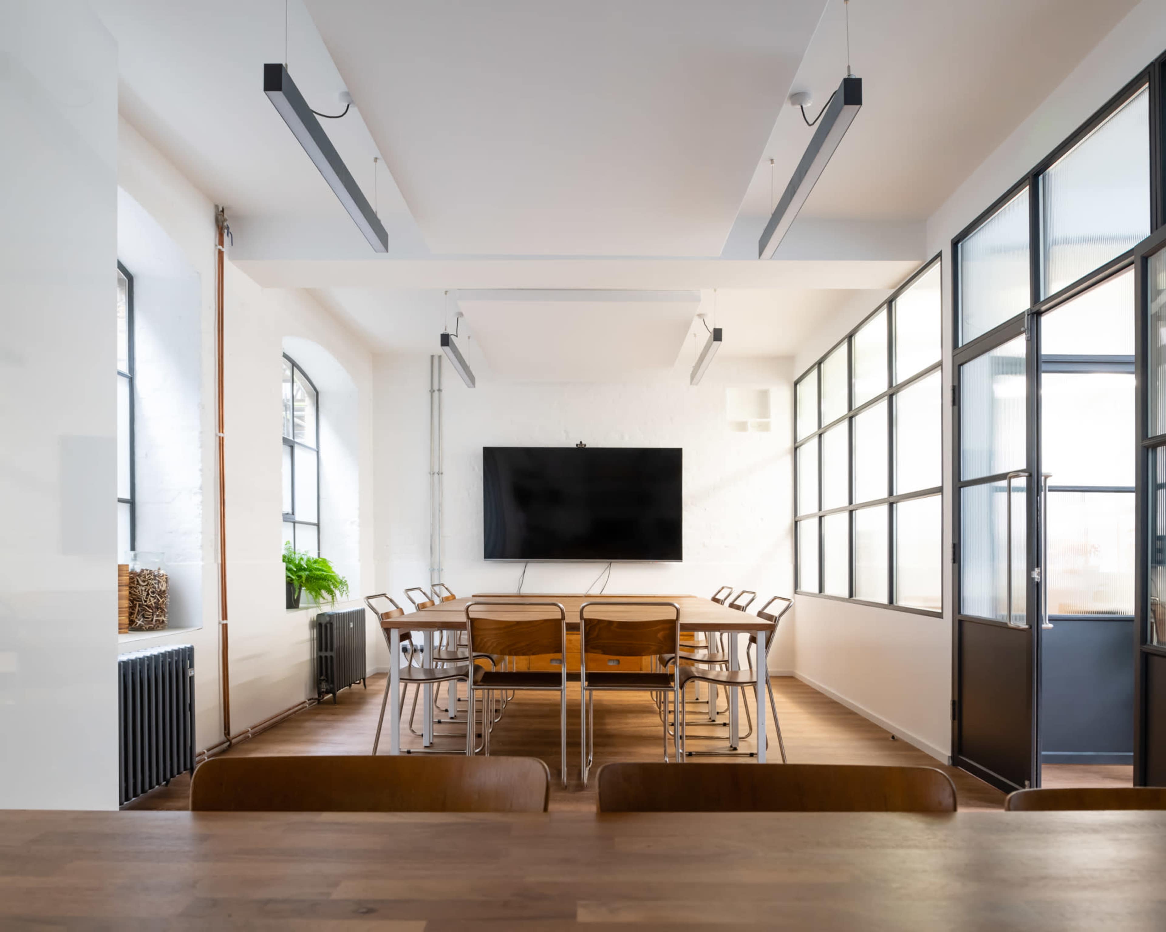 A modern conference room features a long wooden table surrounded by chairs, with a large television mounted on the wall and large windows providing natural light.