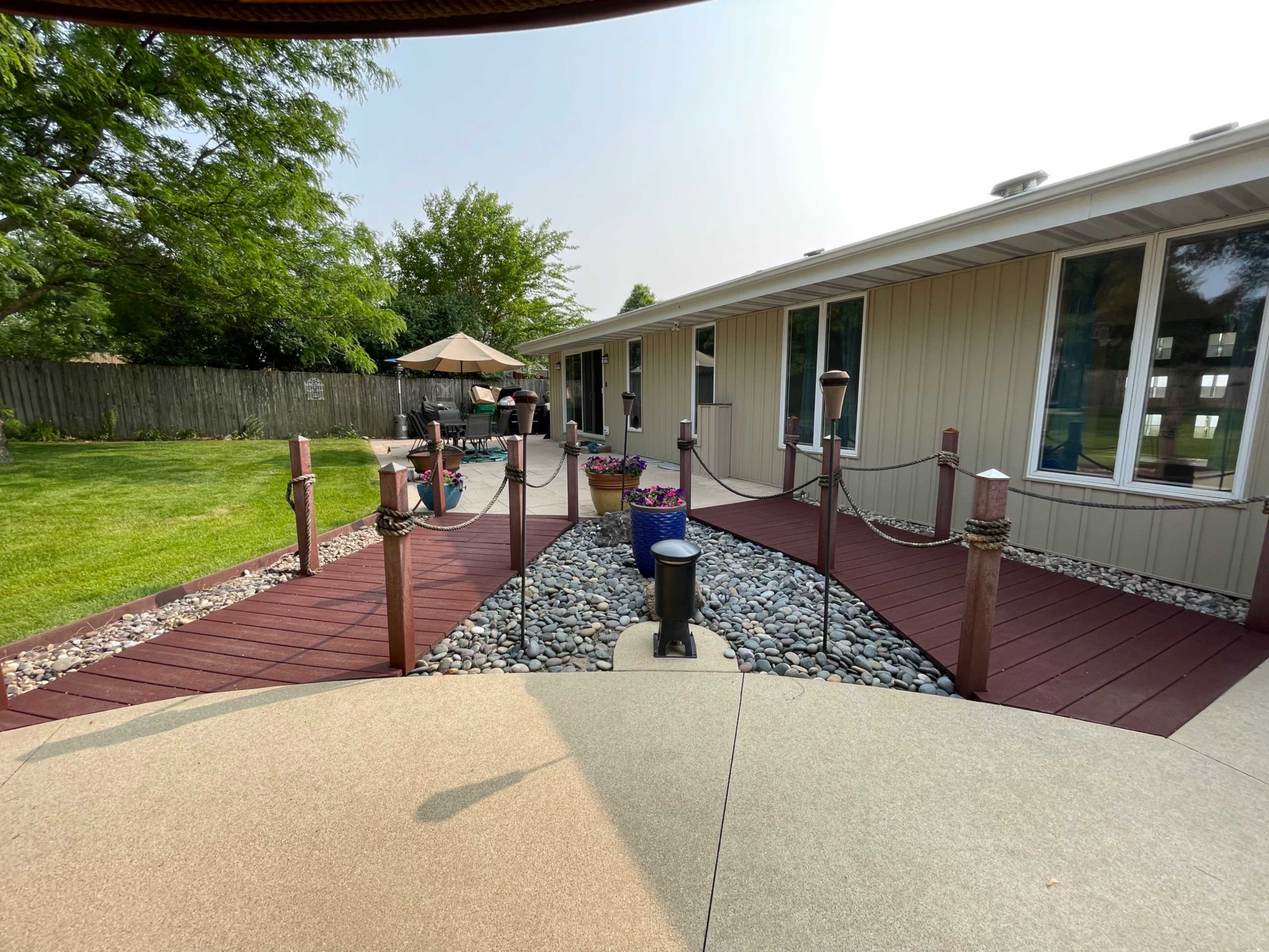 A landscaped backyard features a pathway bordered by stones and plants, leading to a house with a patio and outdoor seating area.