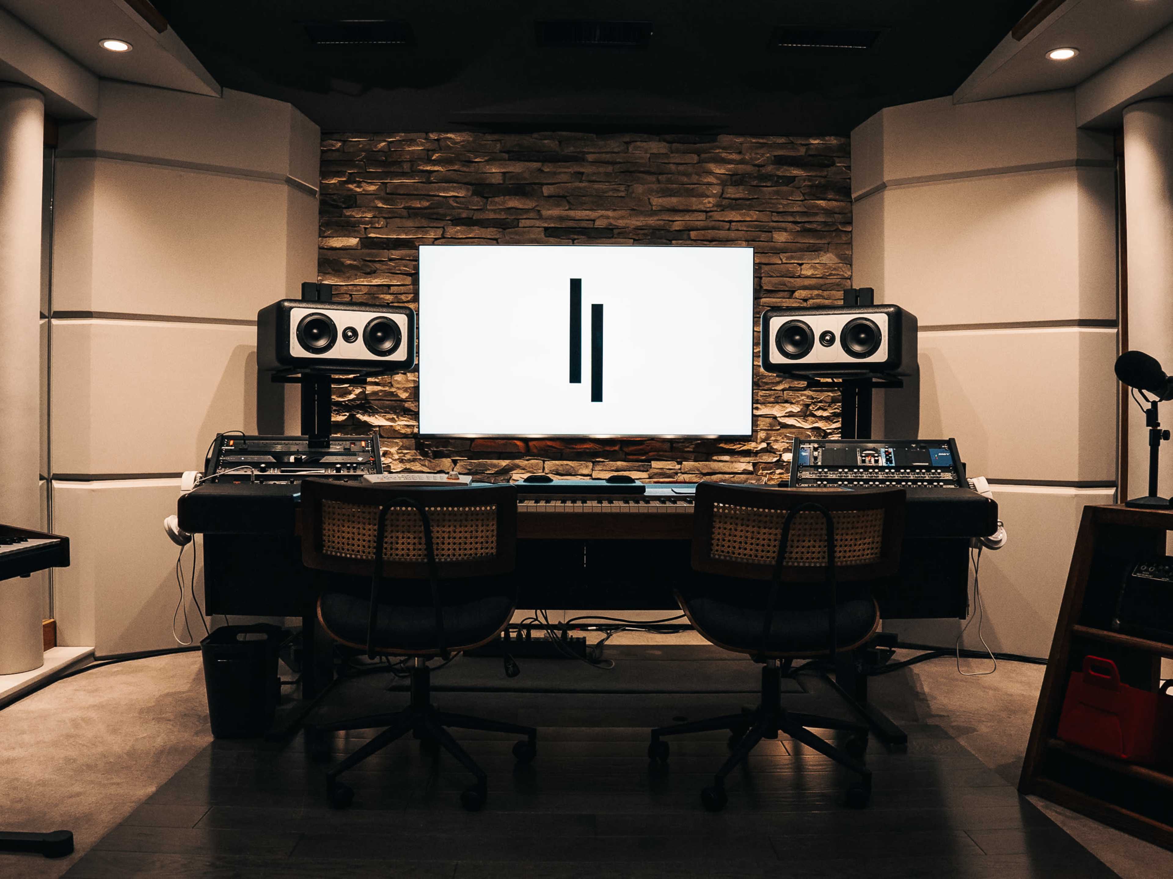 The image shows a modern music studio featuring a central desk with two chairs, a keyboard, audio equipment, and large speakers positioned against a stone wall with a screen displaying a logo.