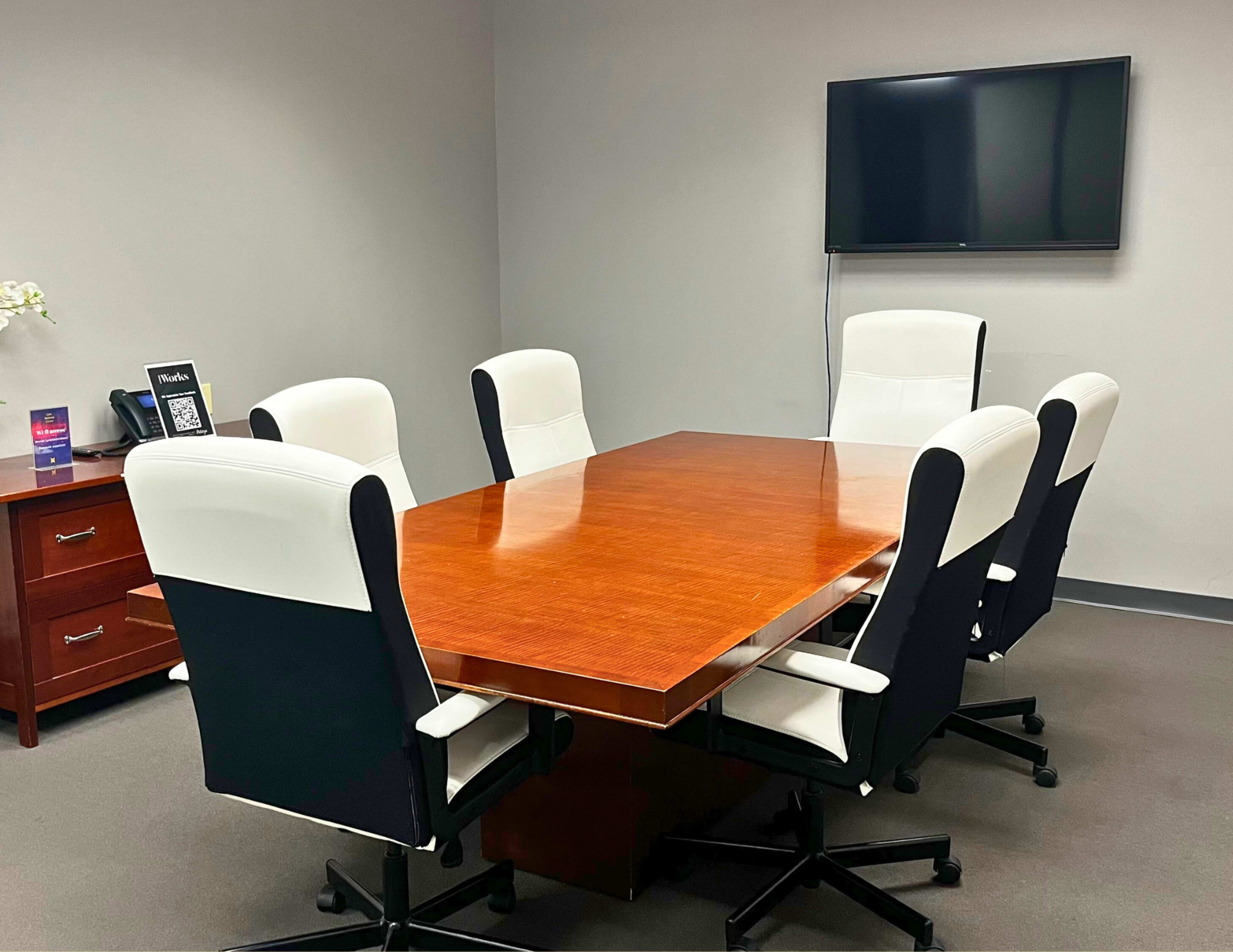 A conference room features a wooden table surrounded by six white and black office chairs, with a television mounted on the wall.