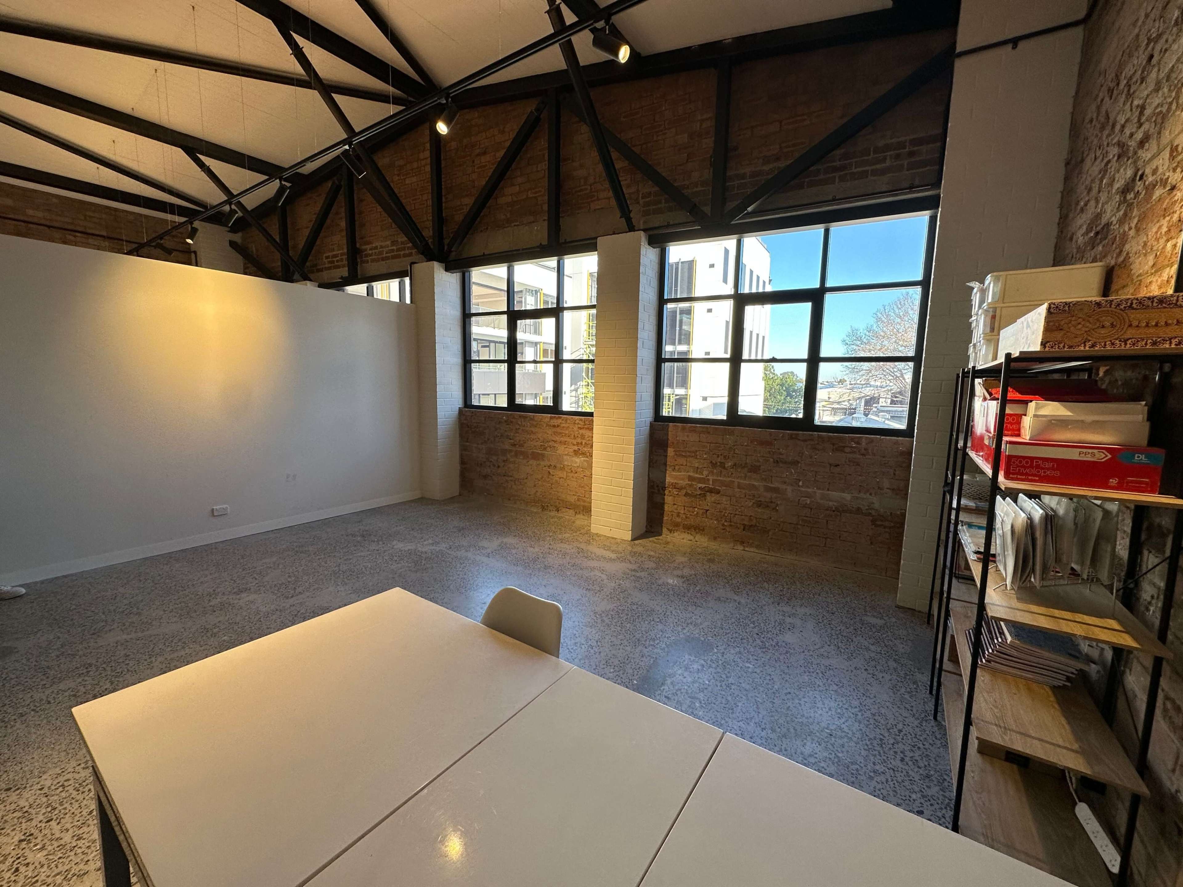 An empty room with exposed brick walls, large windows, and minimal furniture including a table and chairs.