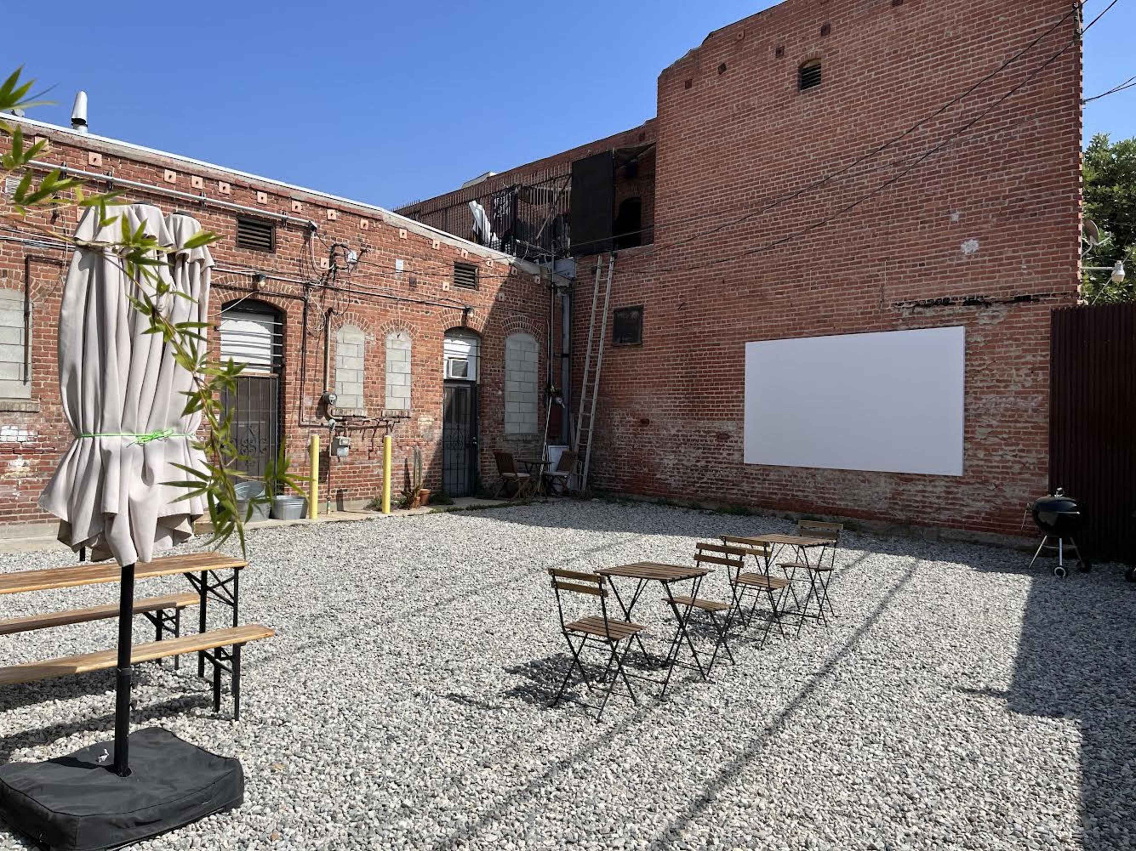 The image shows a gravel courtyard in front of a brick wall, featuring several small tables and chairs, a large white screen, and a BBQ grill under a shaded umbrella.