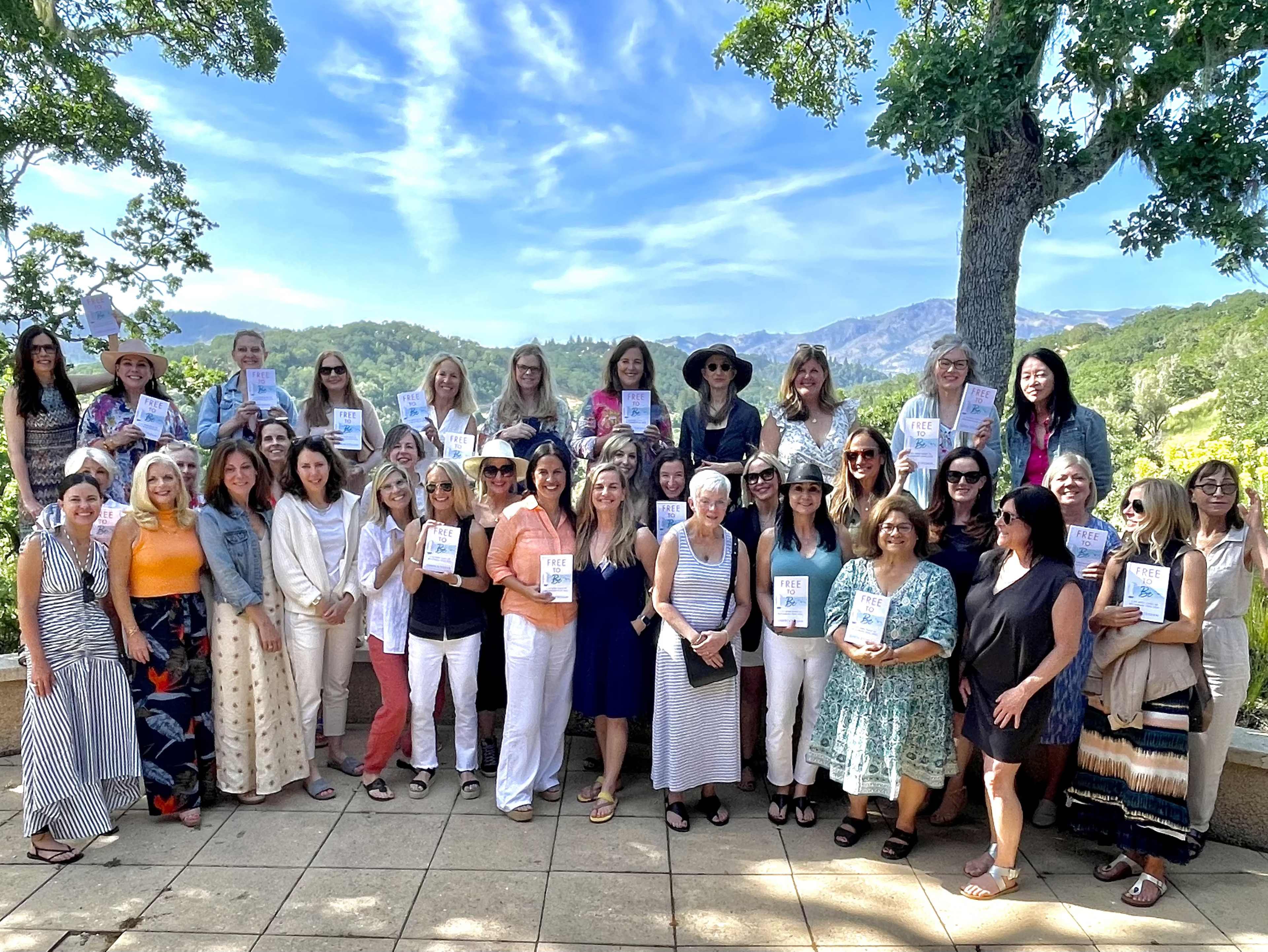 A large group of women stands together outdoors on a patio, holding book copies with a scenic view of mountains and trees in the background.