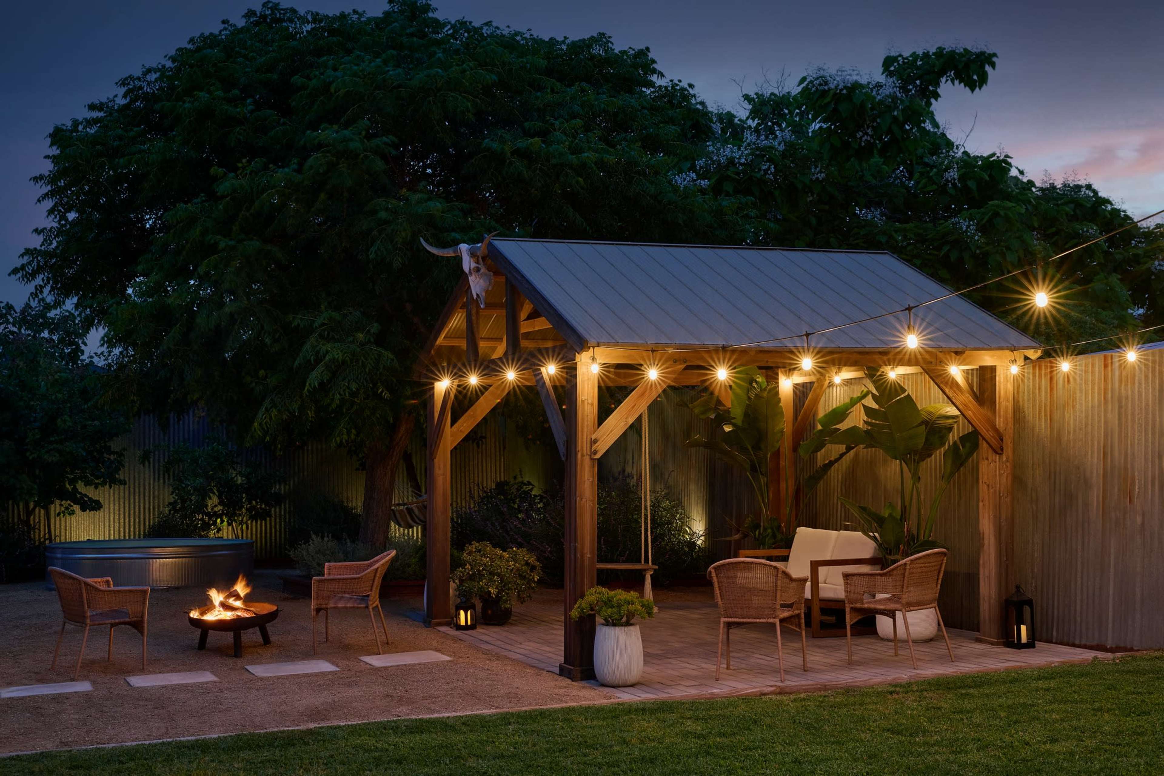 A wooden pavilion with string lights is set up in a landscaped backyard, featuring seating areas around a fire pit.