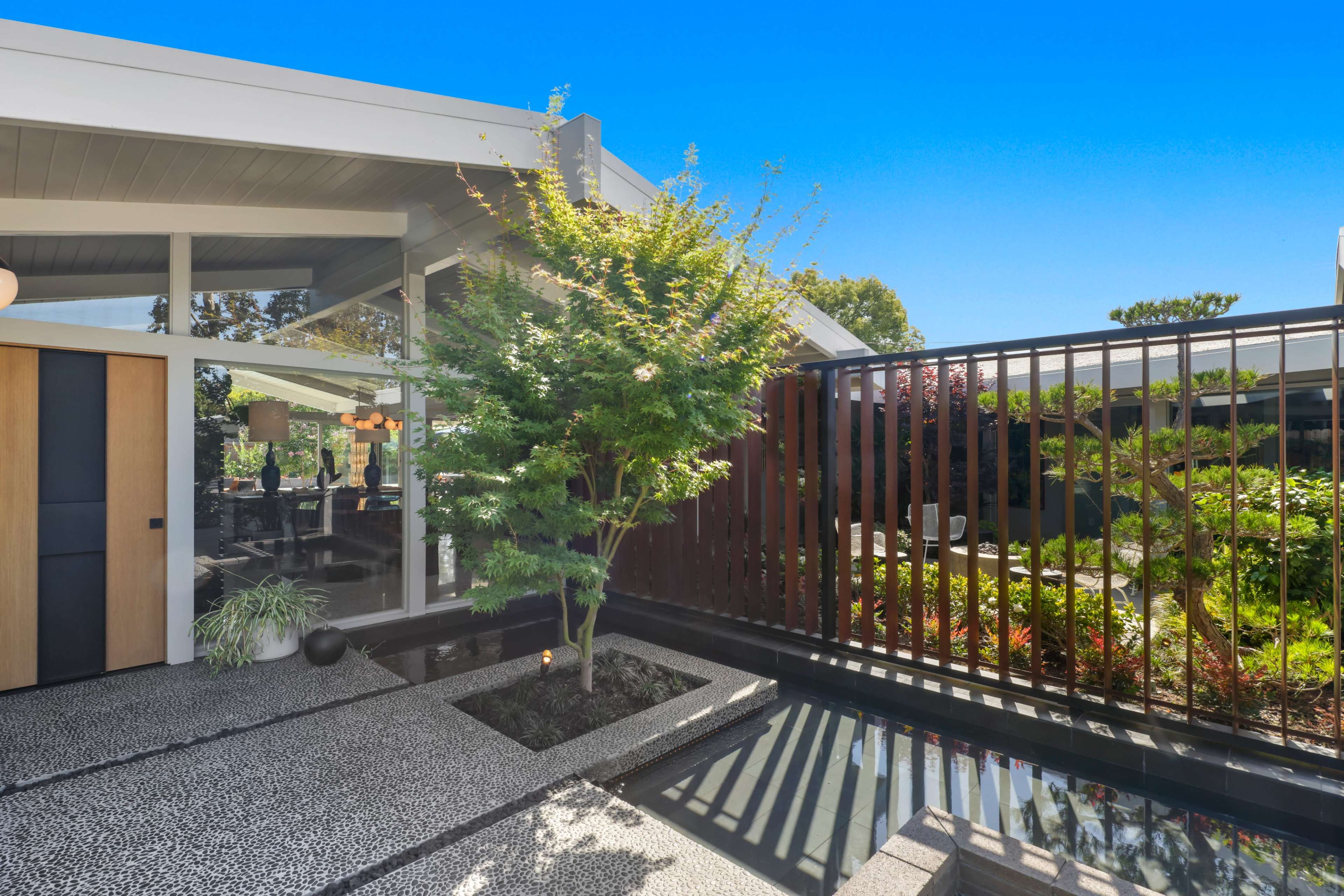 A modern home exterior featuring a landscaped courtyard with a small tree, decorative stone pathways, and a reflective water feature.