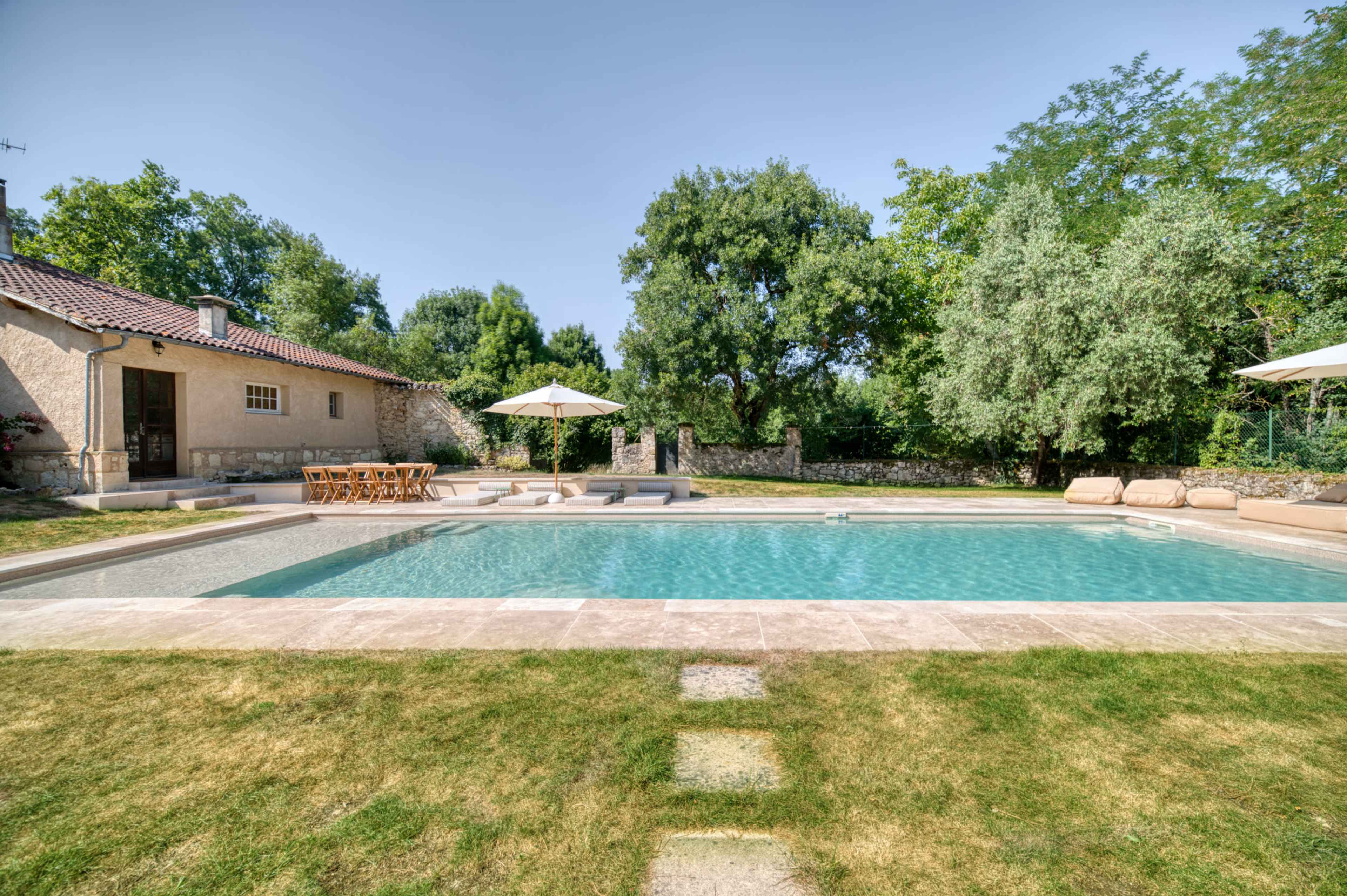 The image shows a spacious backyard featuring a swimming pool, a dining area with a wooden table, and lounge chairs surrounded by lush greenery.
