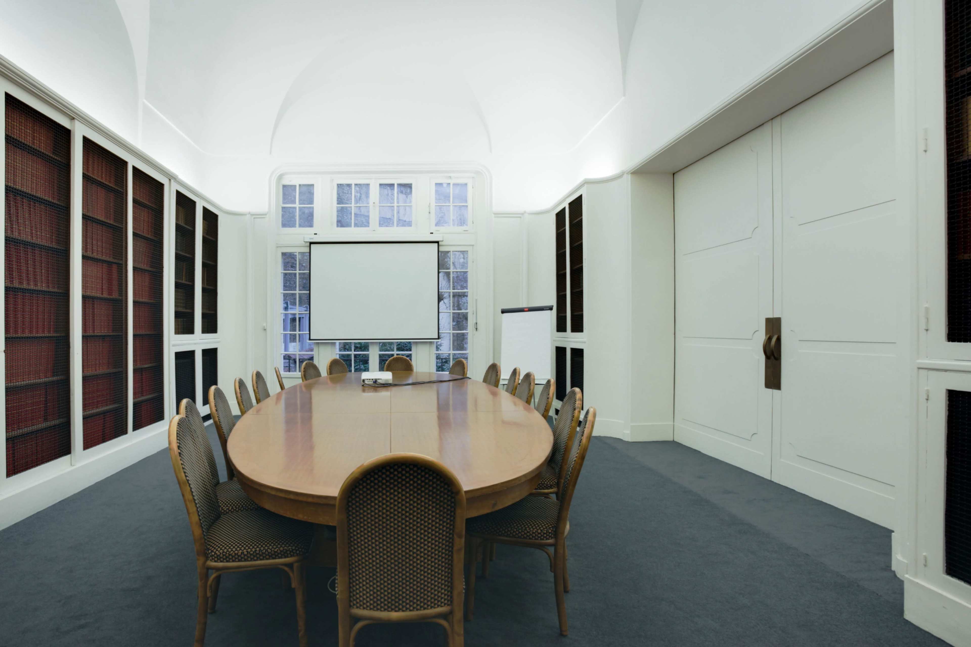 A long oval wooden conference table is surrounded by chairs in a well-lit meeting room with large windows and a blank projection screen at one end.