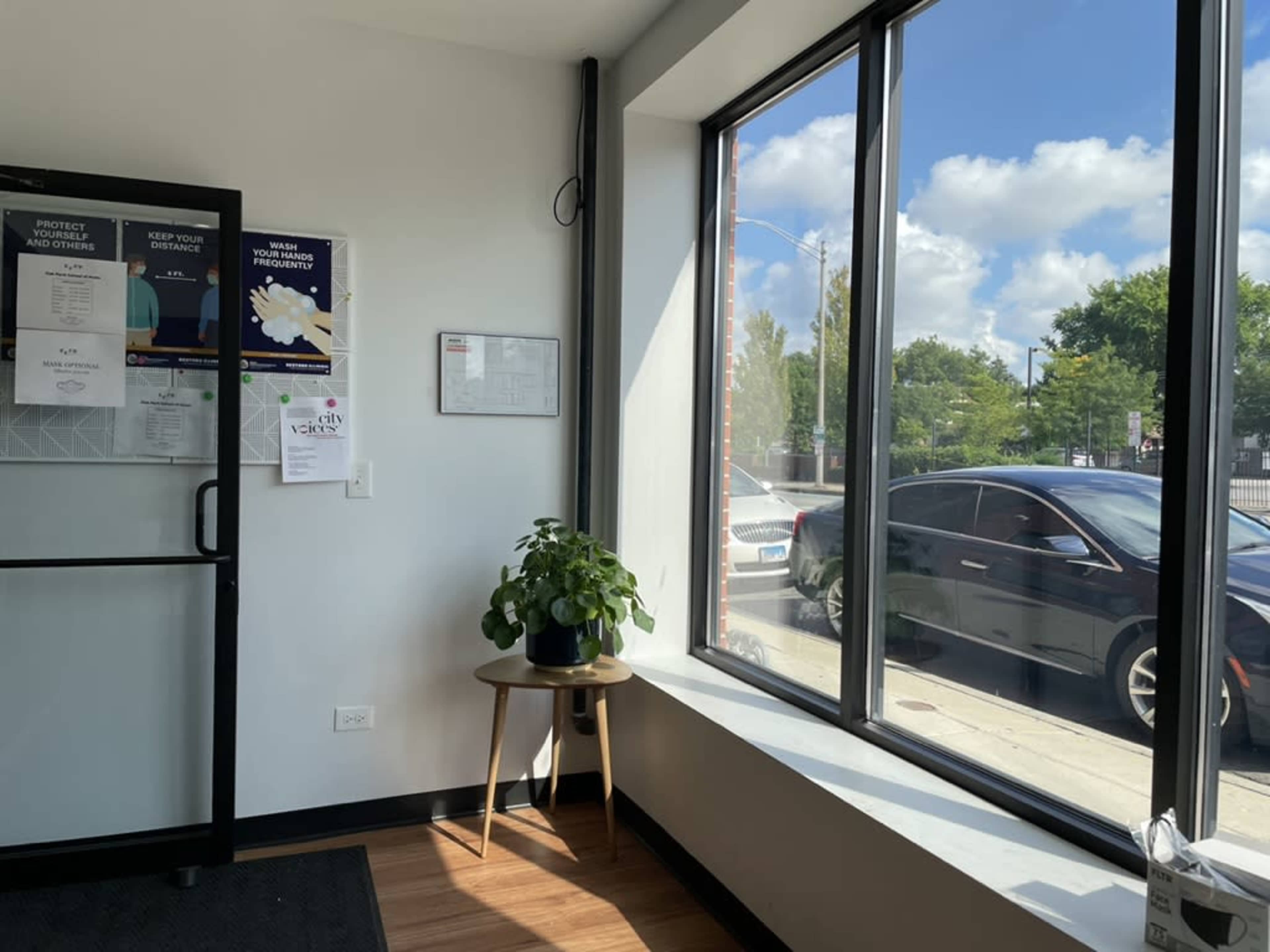 The image shows a corner of a waiting area with large windows, a potted plant on a small table, and a view of a parked car outside.