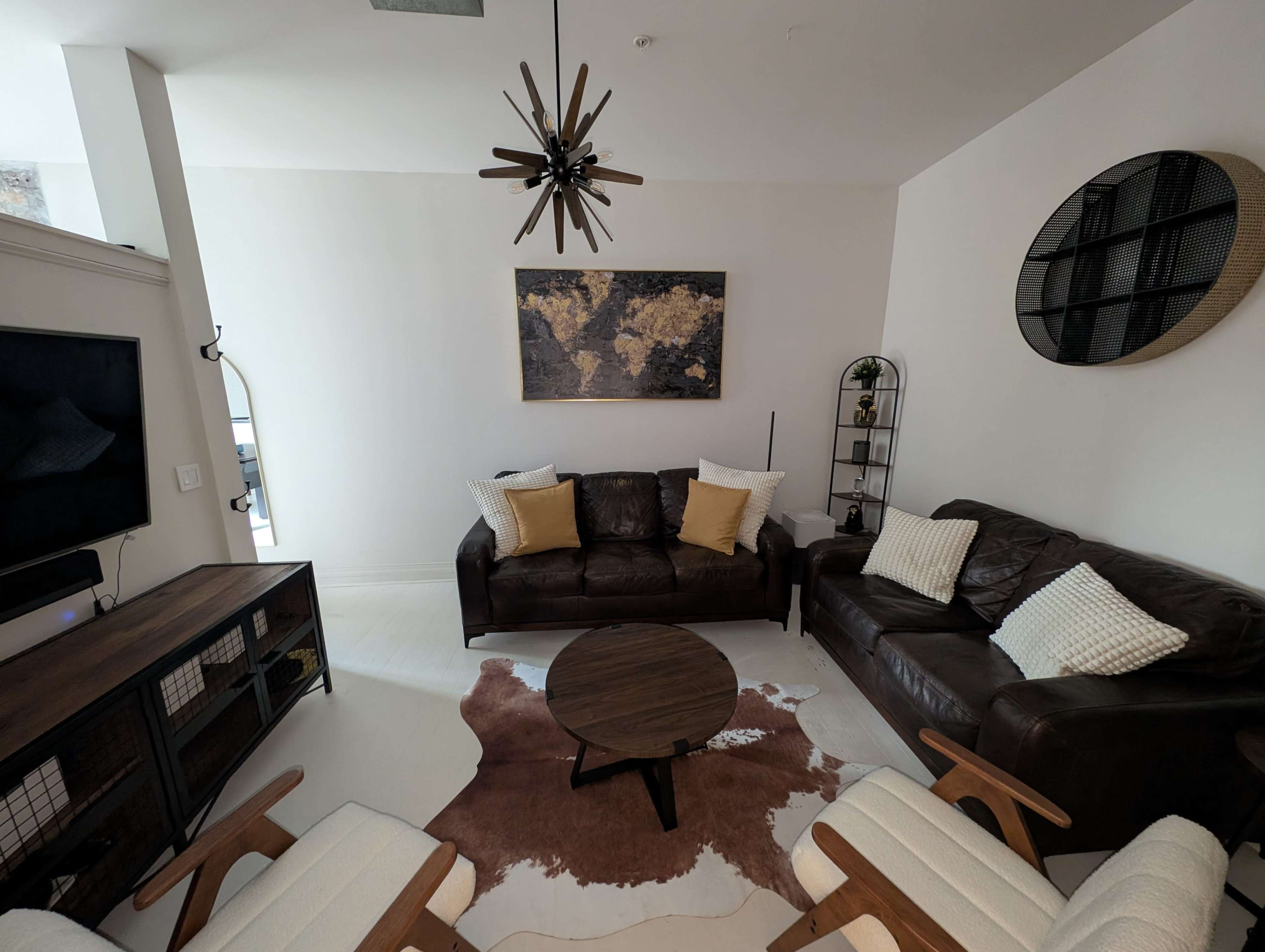 The image shows a modern living room with two brown leather sofas, a round coffee table on a cowhide rug, and a decorative wall art piece.
