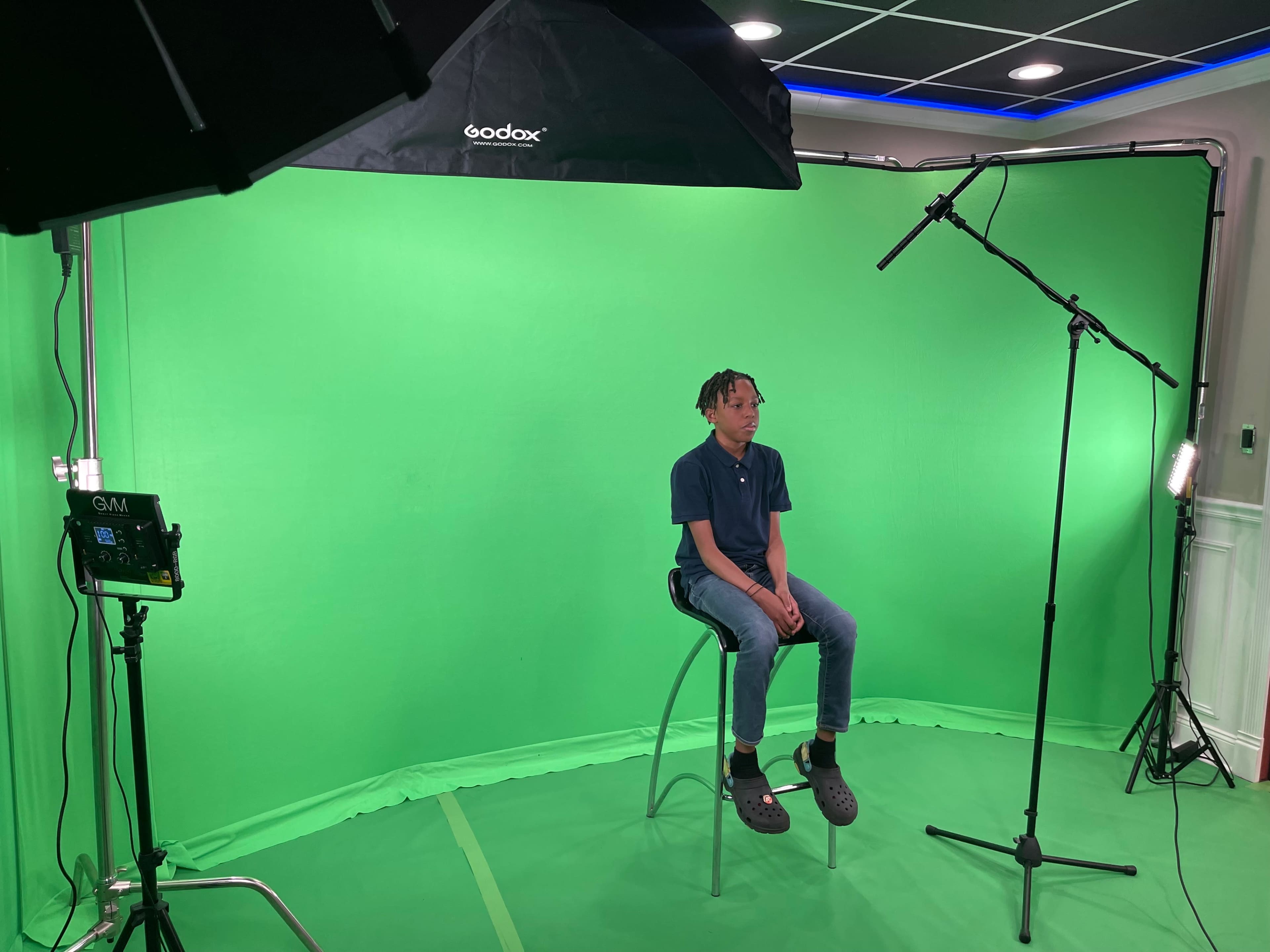 A person sits on a stool in front of a green screen, surrounded by lighting equipment and a microphone.