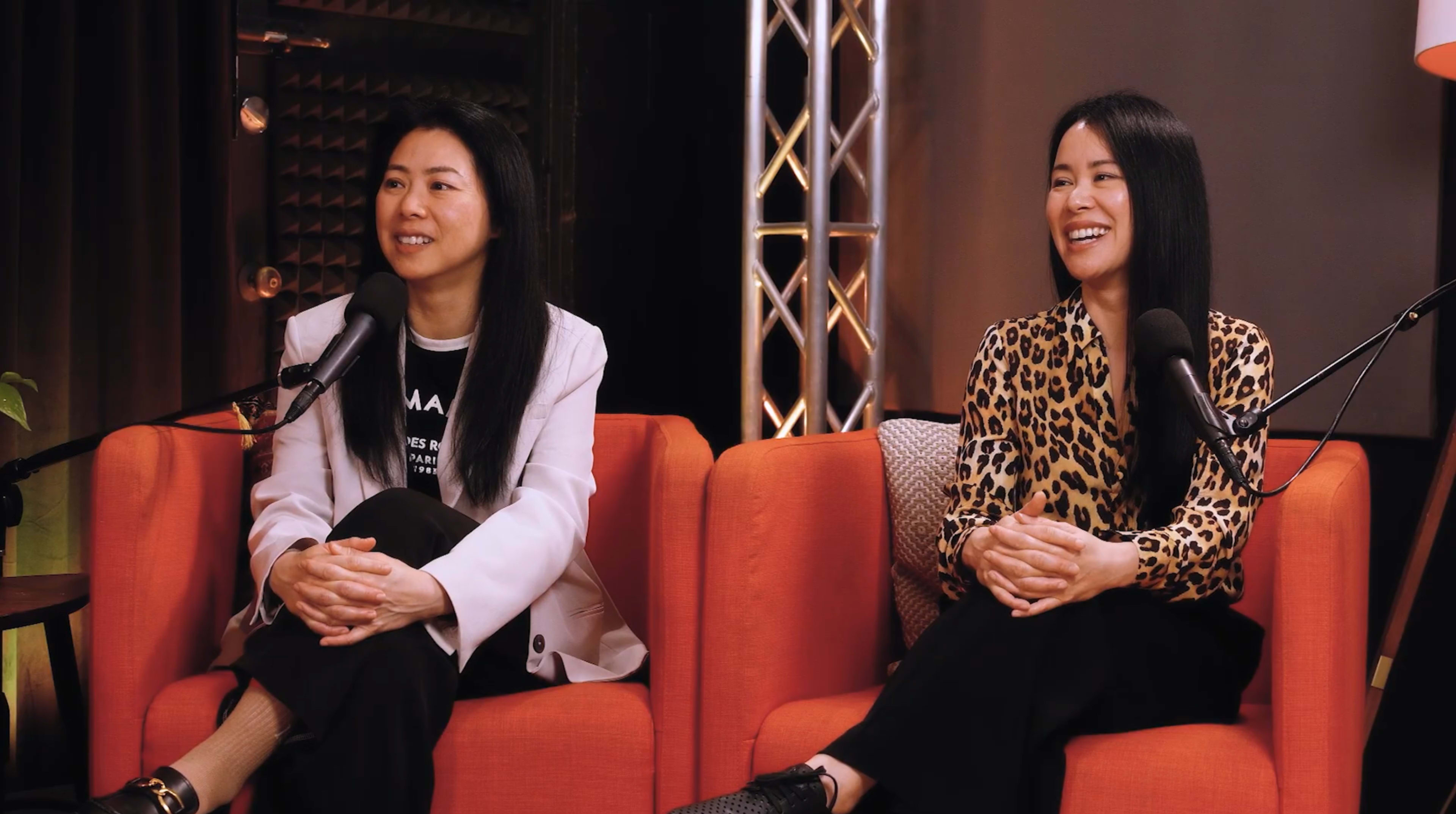Two women sitting on orange couches, engaged in conversation with microphones in front of them.
