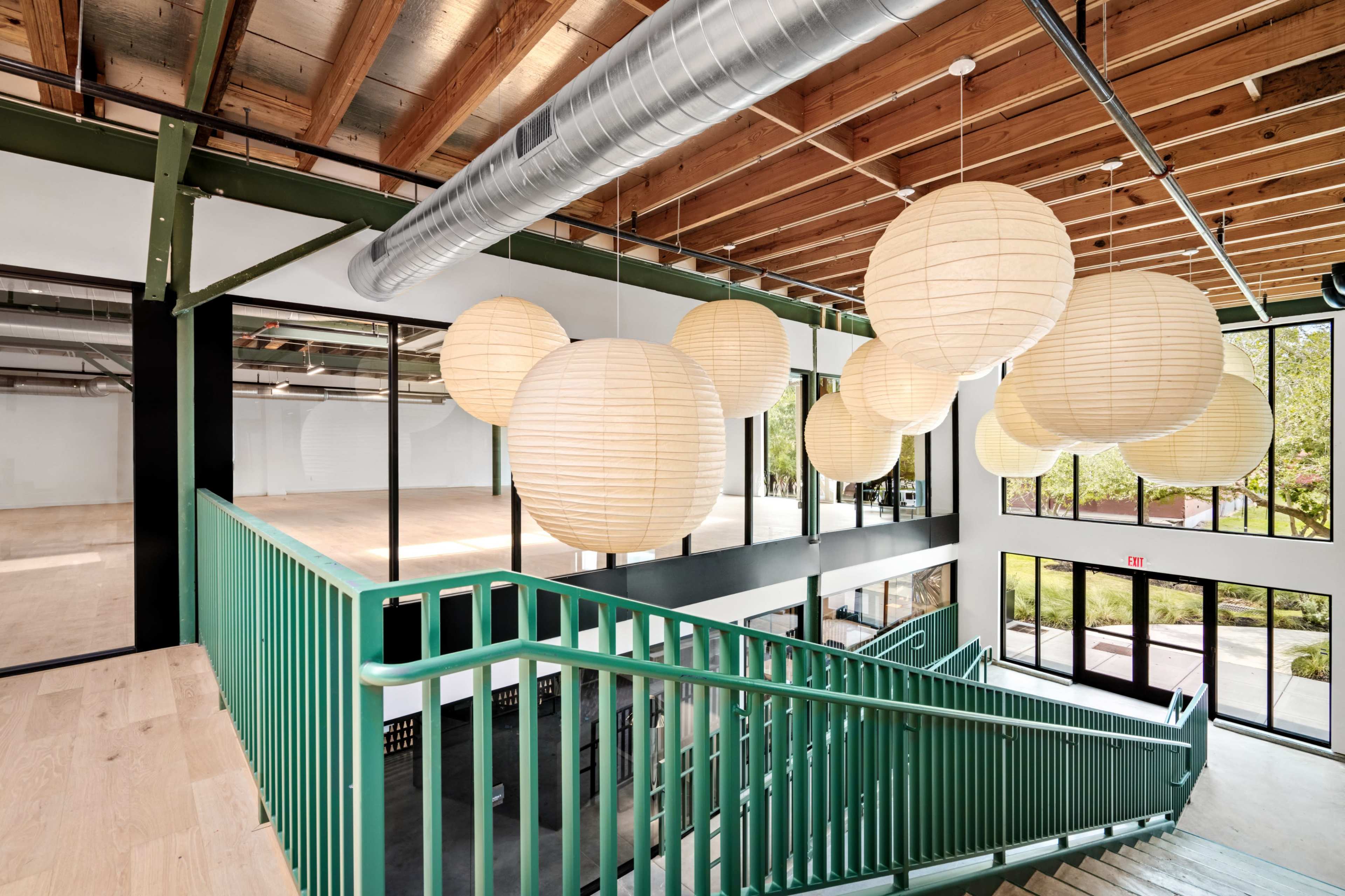 The image shows a modern interior with a staircase, green railings, and large paper lanterns suspended from the ceiling, giving an open and airy feeling to the space.