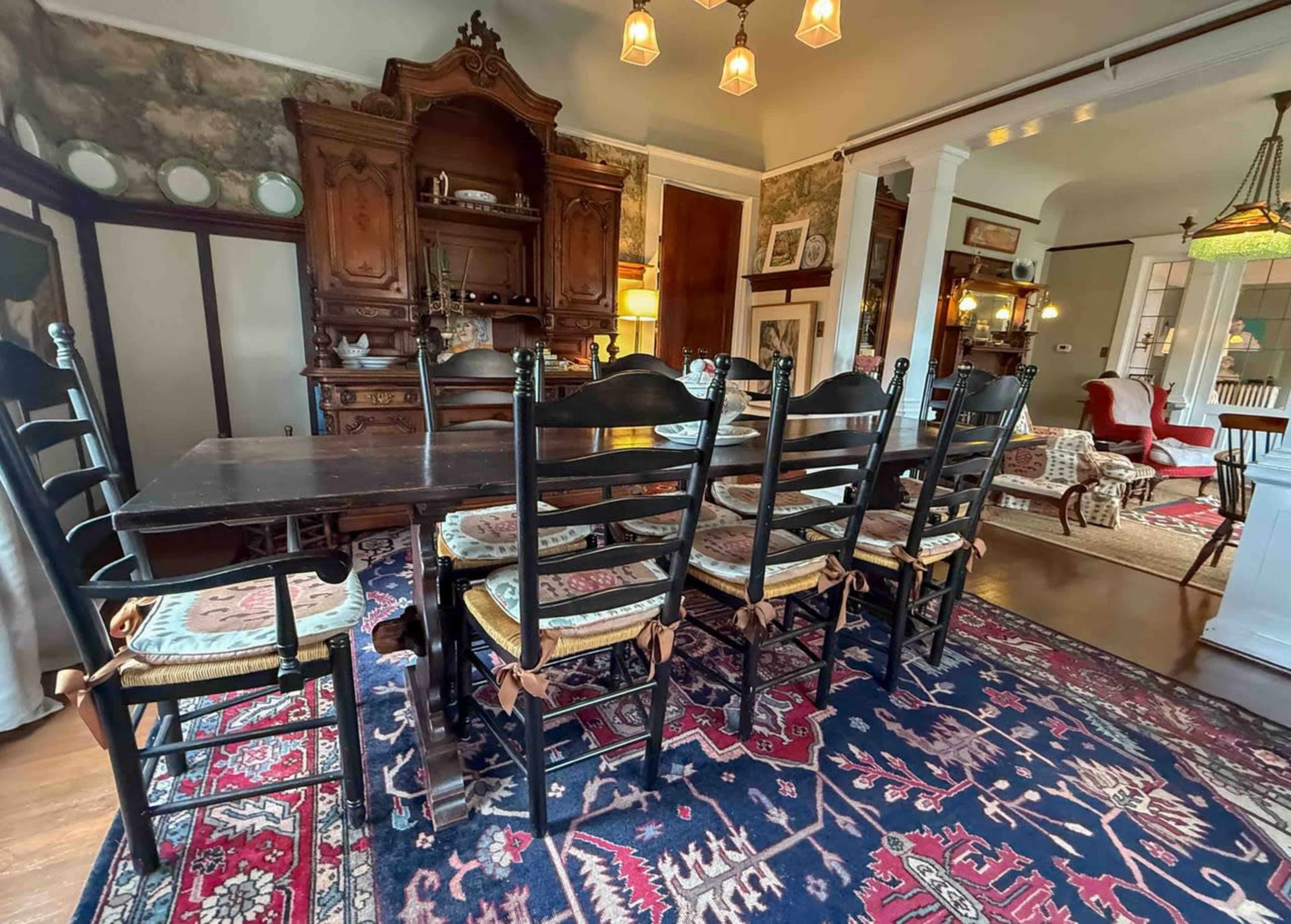 A long wooden dining table with black chairs is set in a room containing vintage furniture and a patterned area rug.