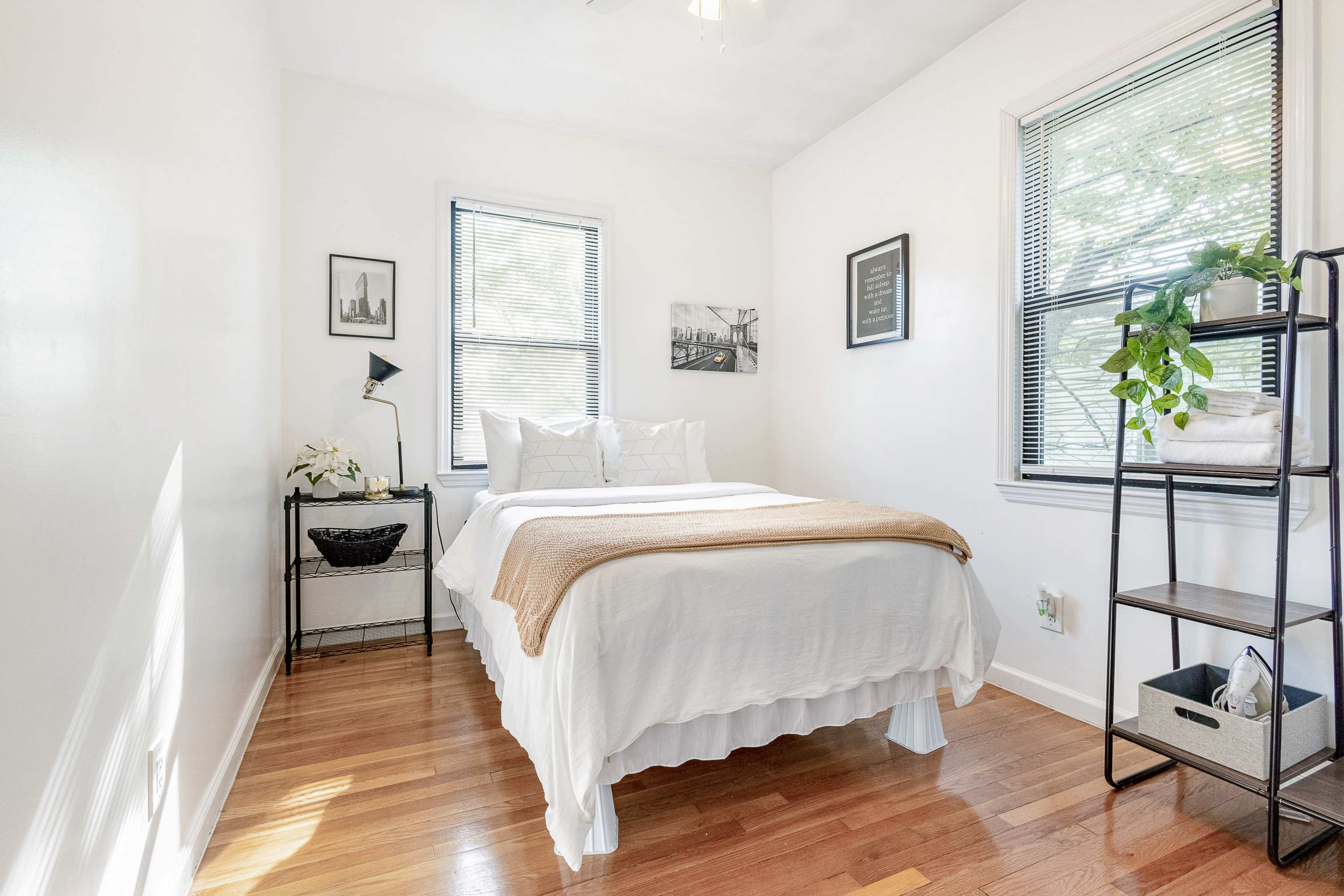 A small bedroom with a bed, two windows, a bedside table, and a shelf with plants and decorative items.