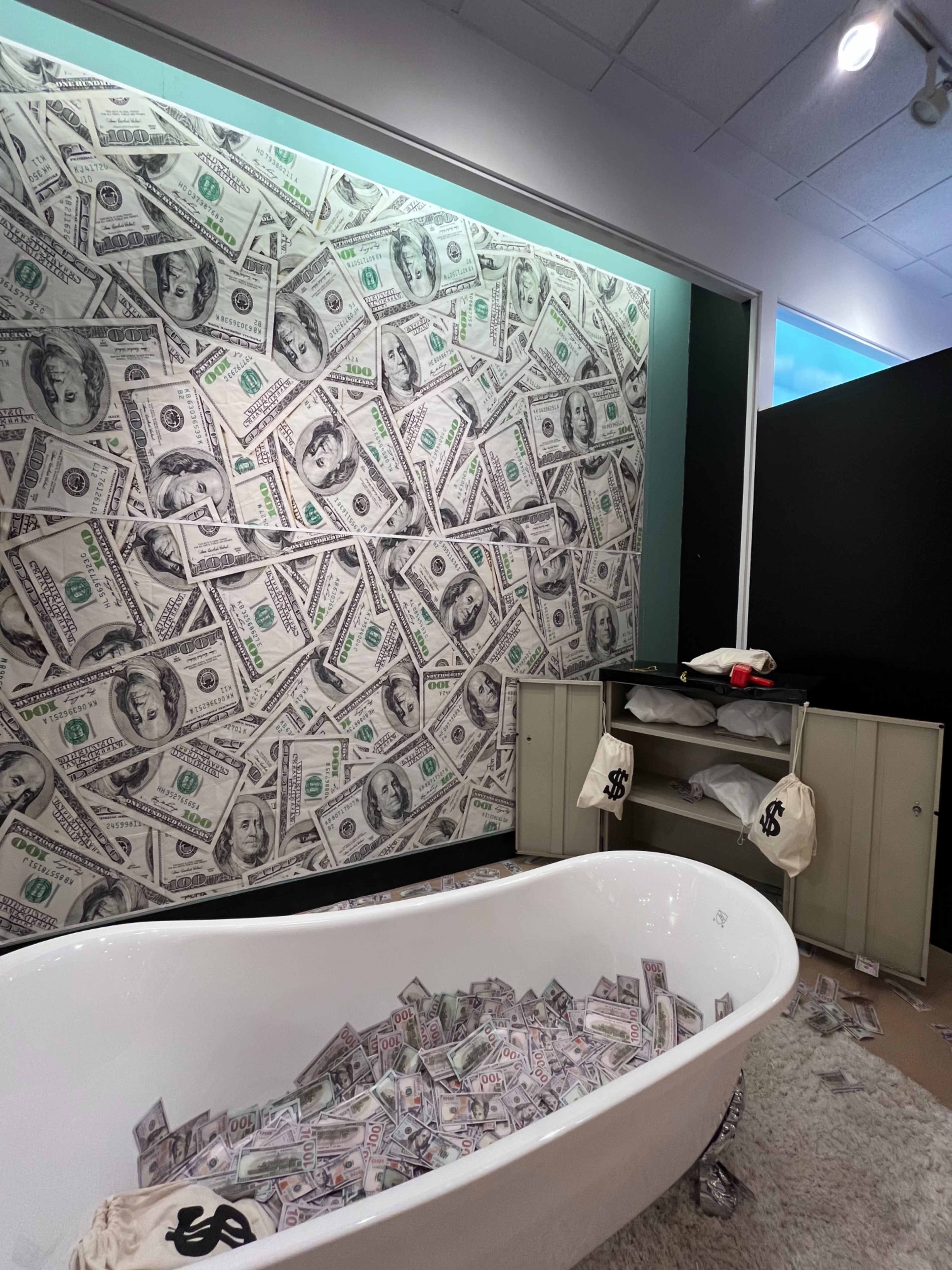 A white bathtub filled with what appears to be fake money sits in a room with a wall covered in a dollar bill design.