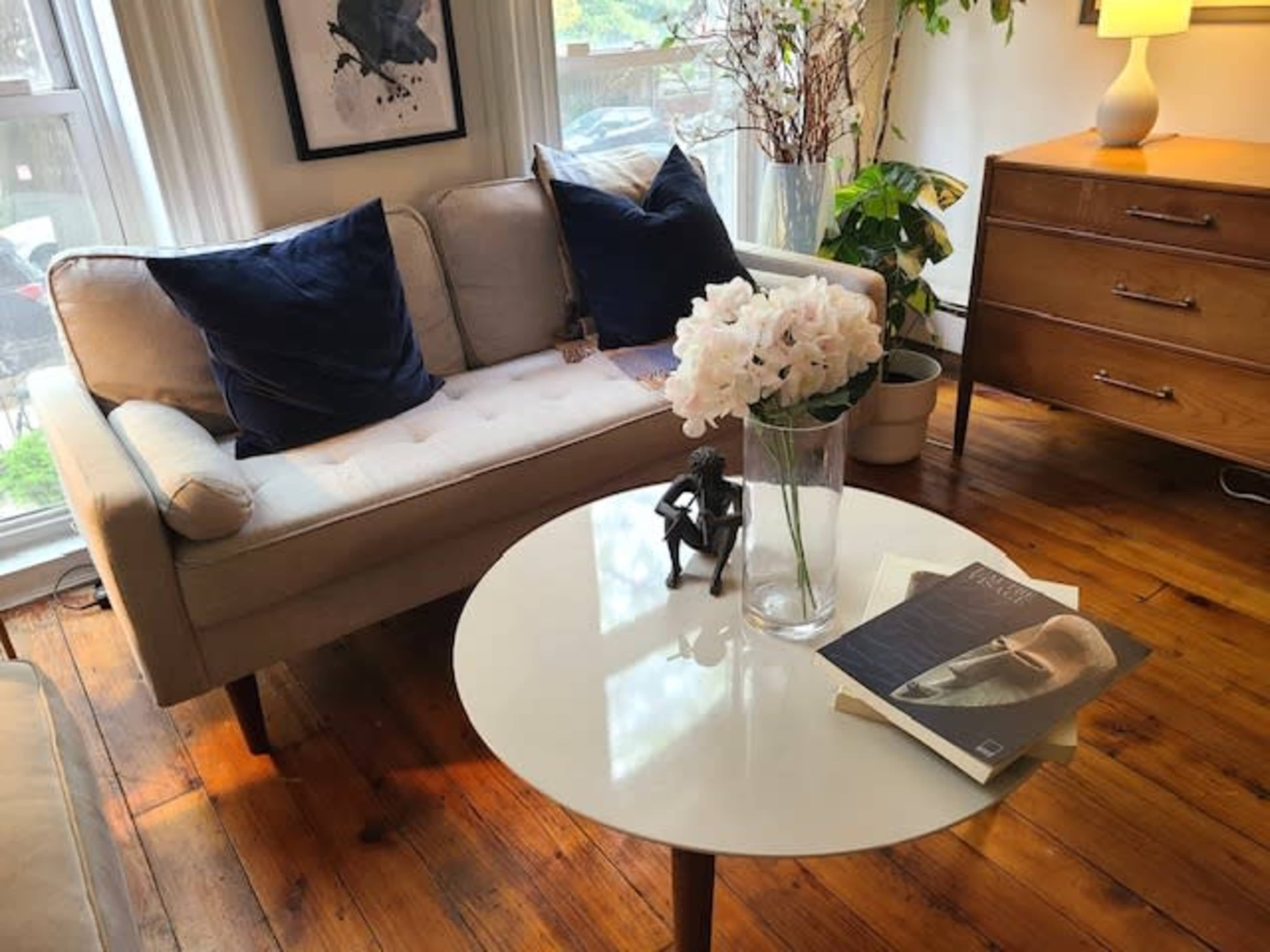 A light-colored sofa with dark cushions sits beside a round coffee table holding a vase of flowers and books, against a backdrop of wooden floors and a large window.