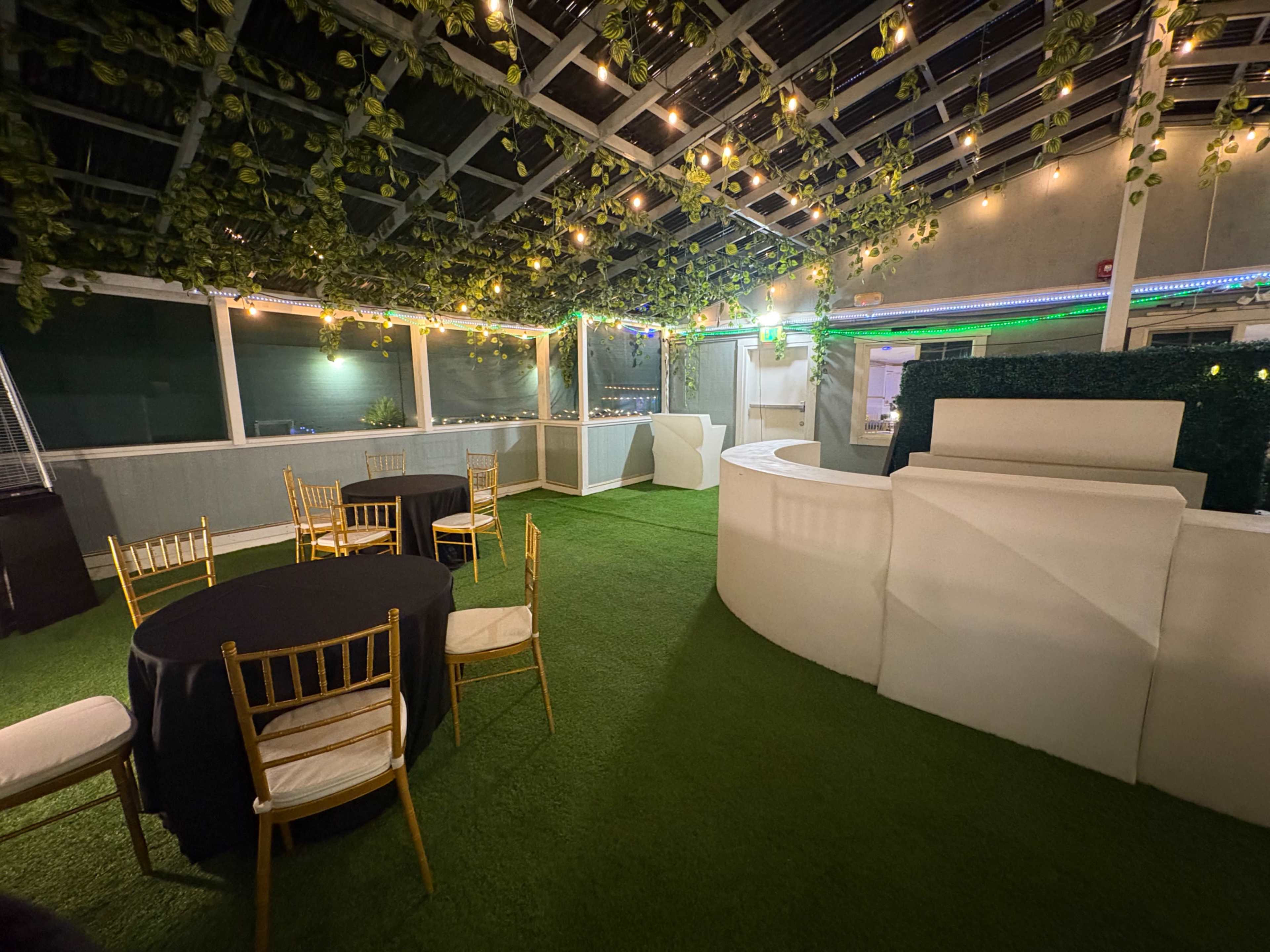 The image shows a partially enclosed event space featuring artificial grass, tables with black and white linens, and overhead string lights surrounded by greenery.