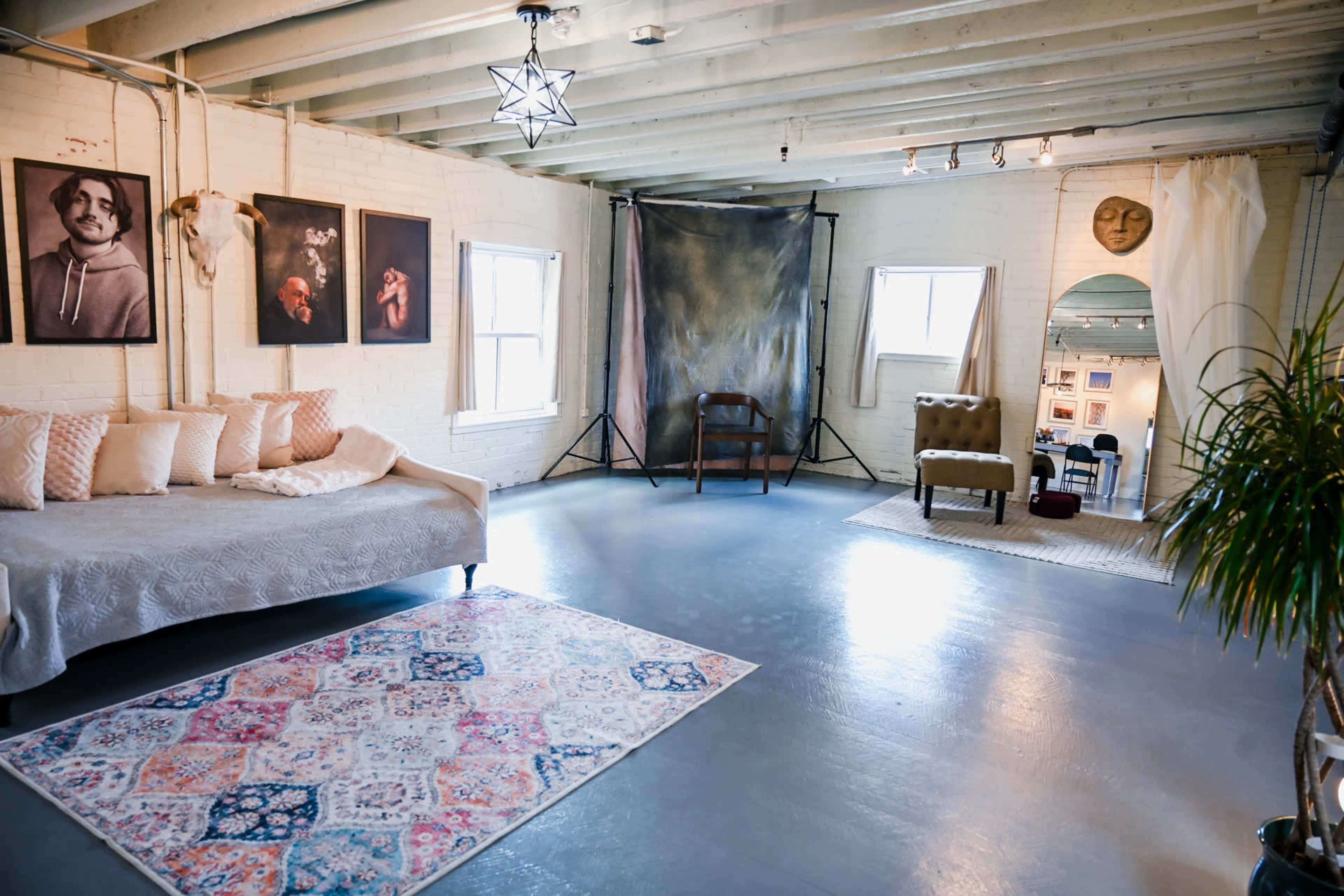 The image shows a spacious studio with a cozy sofa, art on the walls, a rug on the floor, and a set of windows providing natural light.