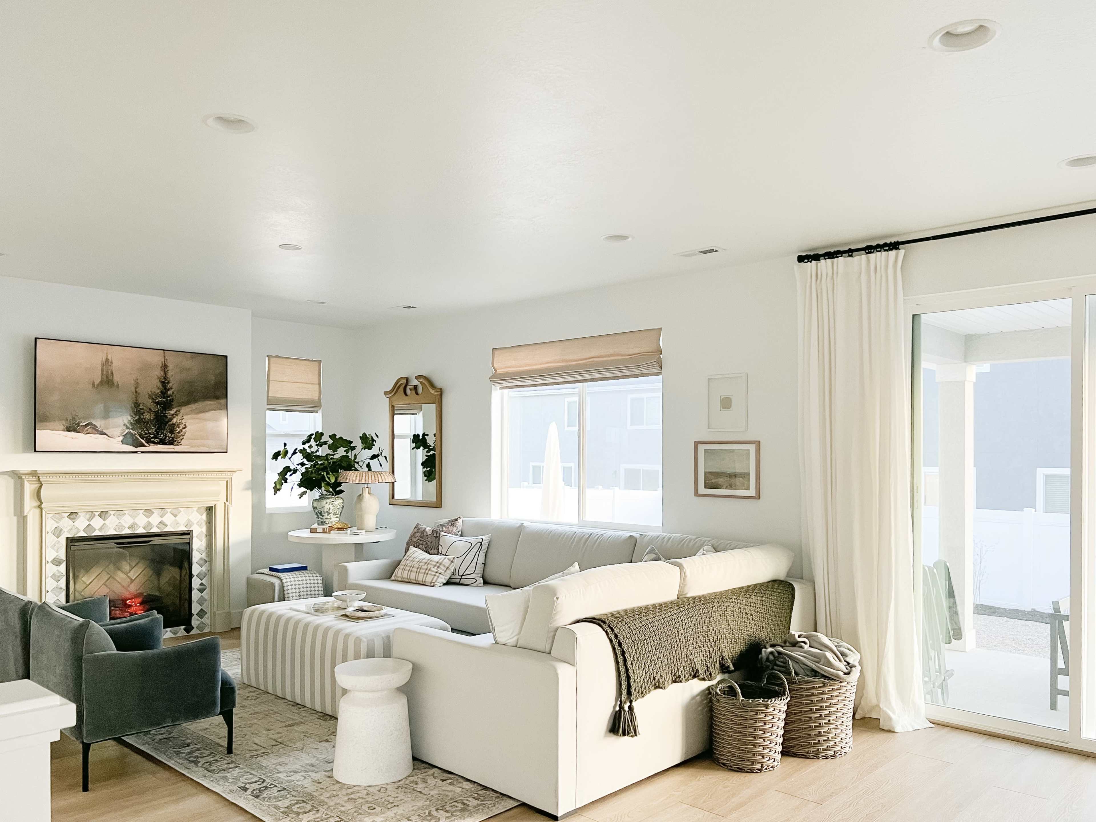 A bright living room features a white sectional sofa, a fireplace with a framed painting above it, and large windows with sheer curtains.