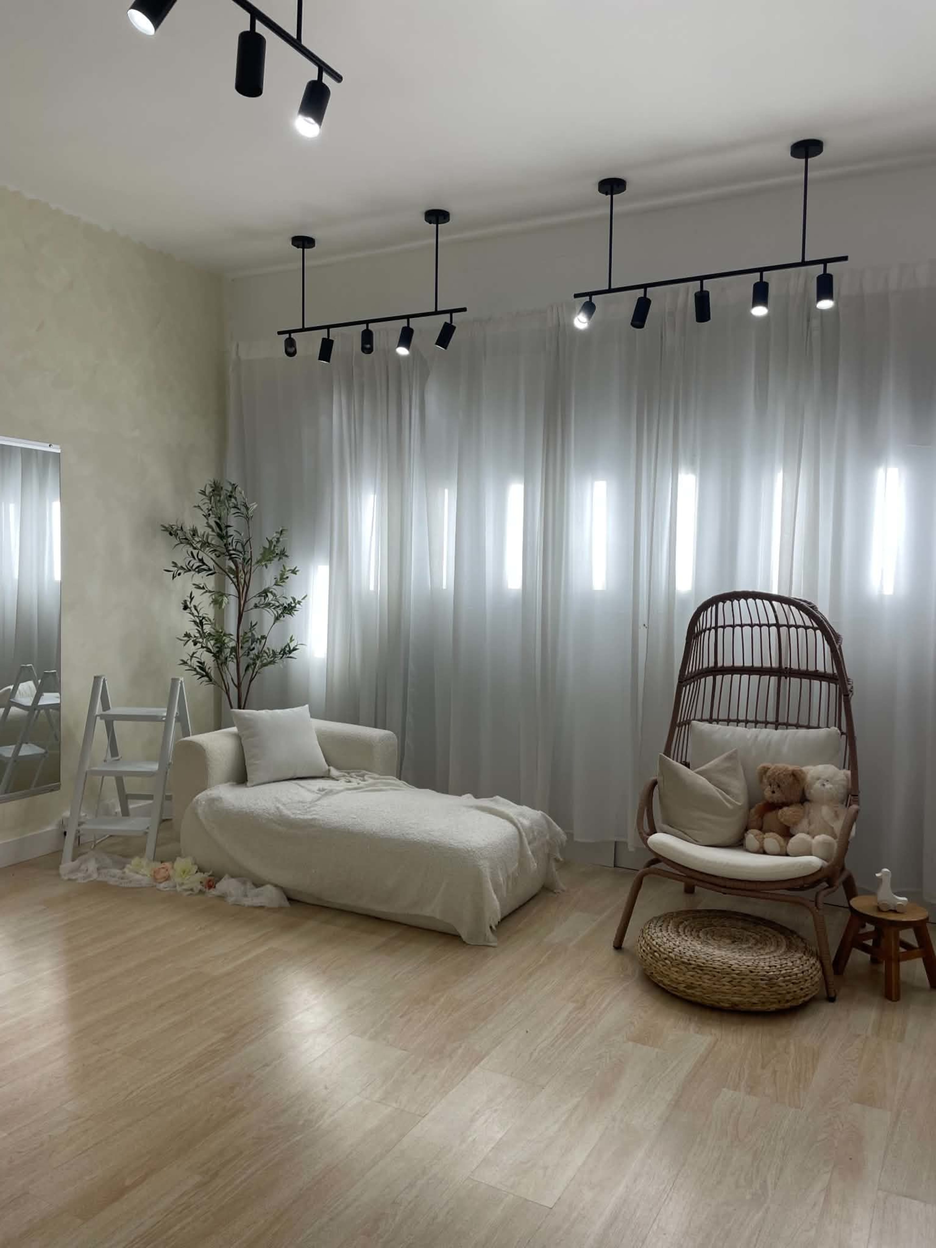 The image shows a light and airy room with a sofa, a decorative ladder, a hanging chair with a teddy bear, and large curtains allowing natural light to filter in.