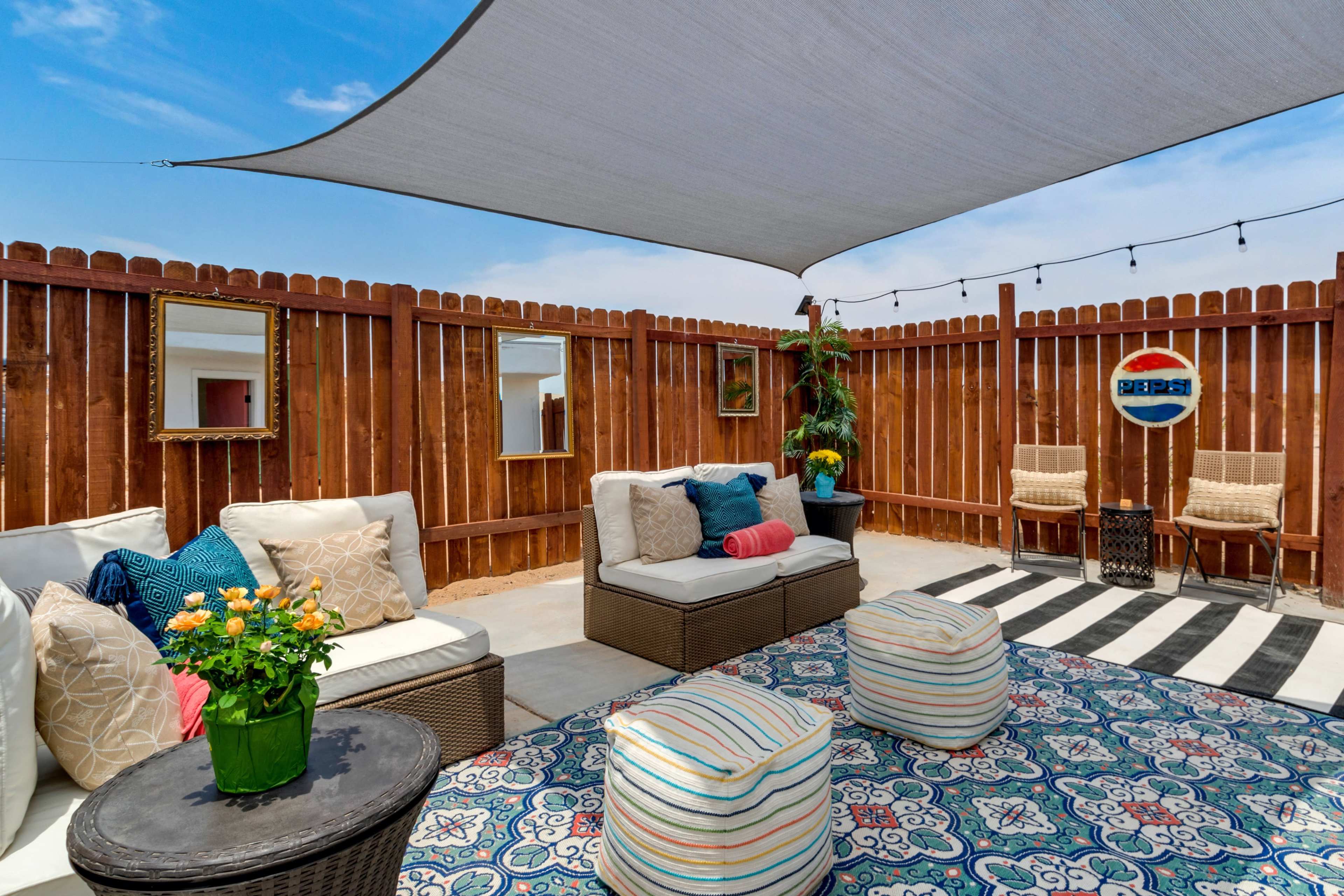 The image showcases an outdoor seating area with two woven chairs, a patterned rug, and a shaded canopy surrounded by a wooden fence.