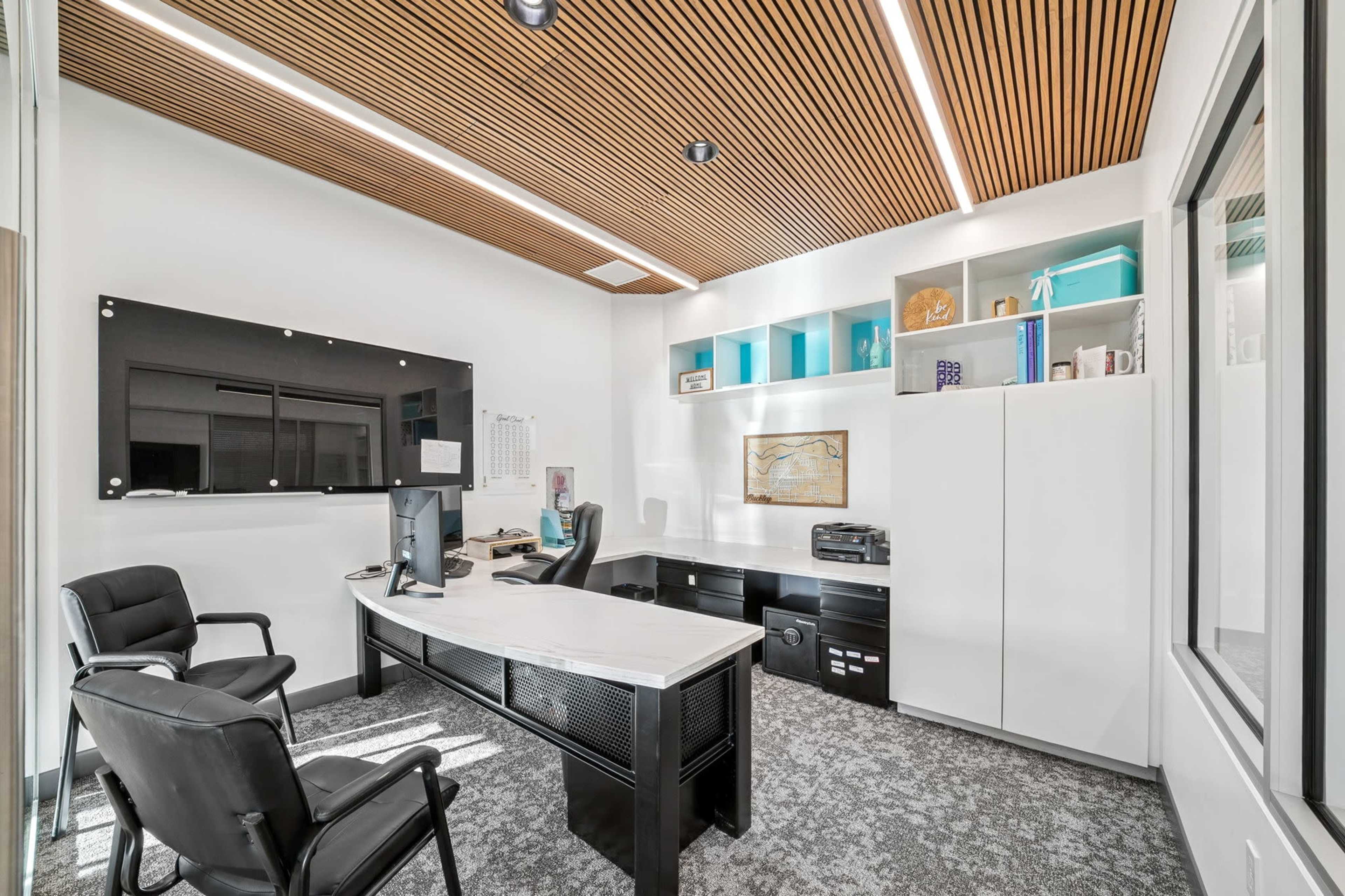 A modern office features a sleek desk, ergonomic chairs, a wall-mounted TV, storage shelves with decorative items, and a large window.