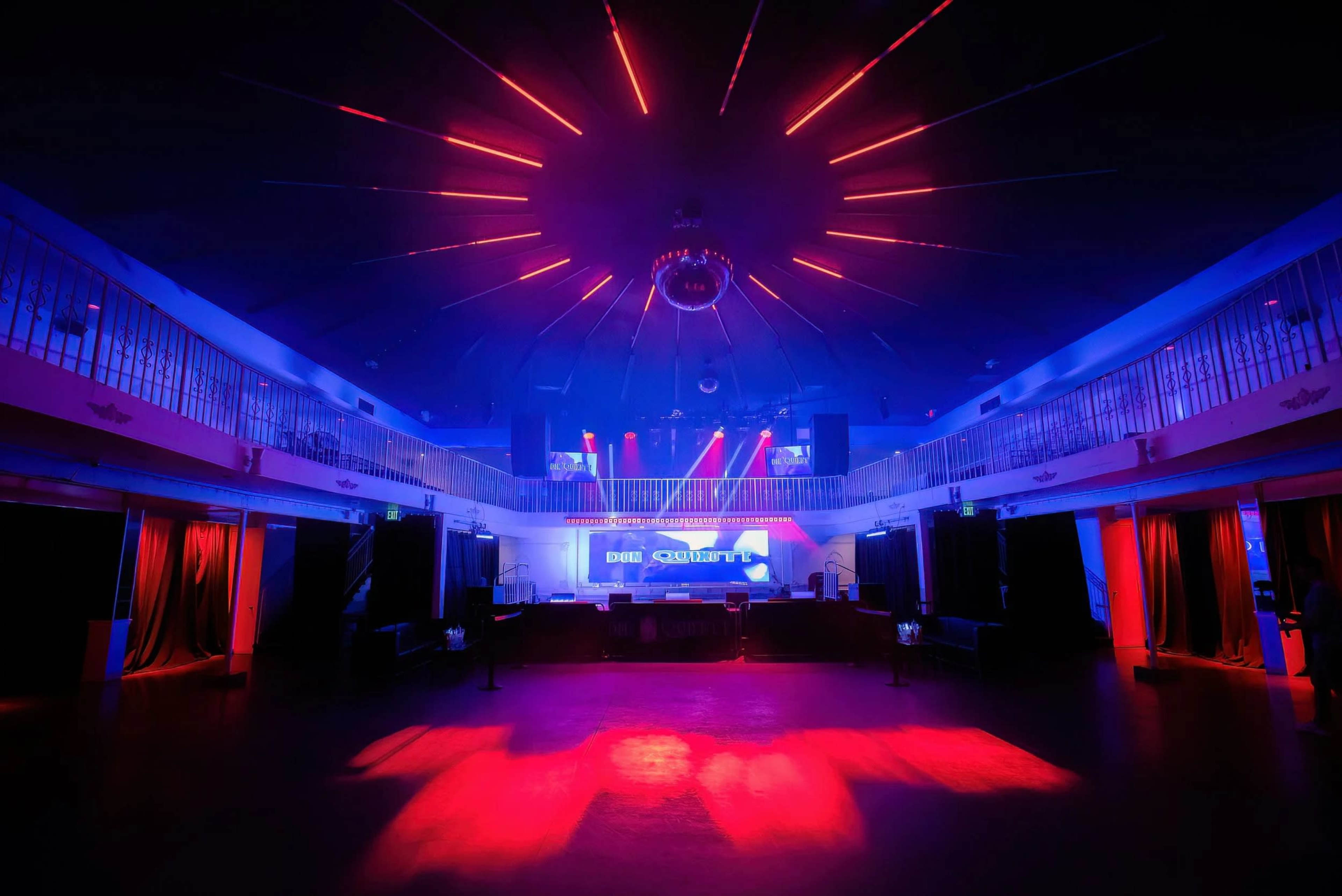 A dimly lit nightclub features vibrant blue and red lighting with a central stage adorned by a disco ball and illuminated by beams of light.