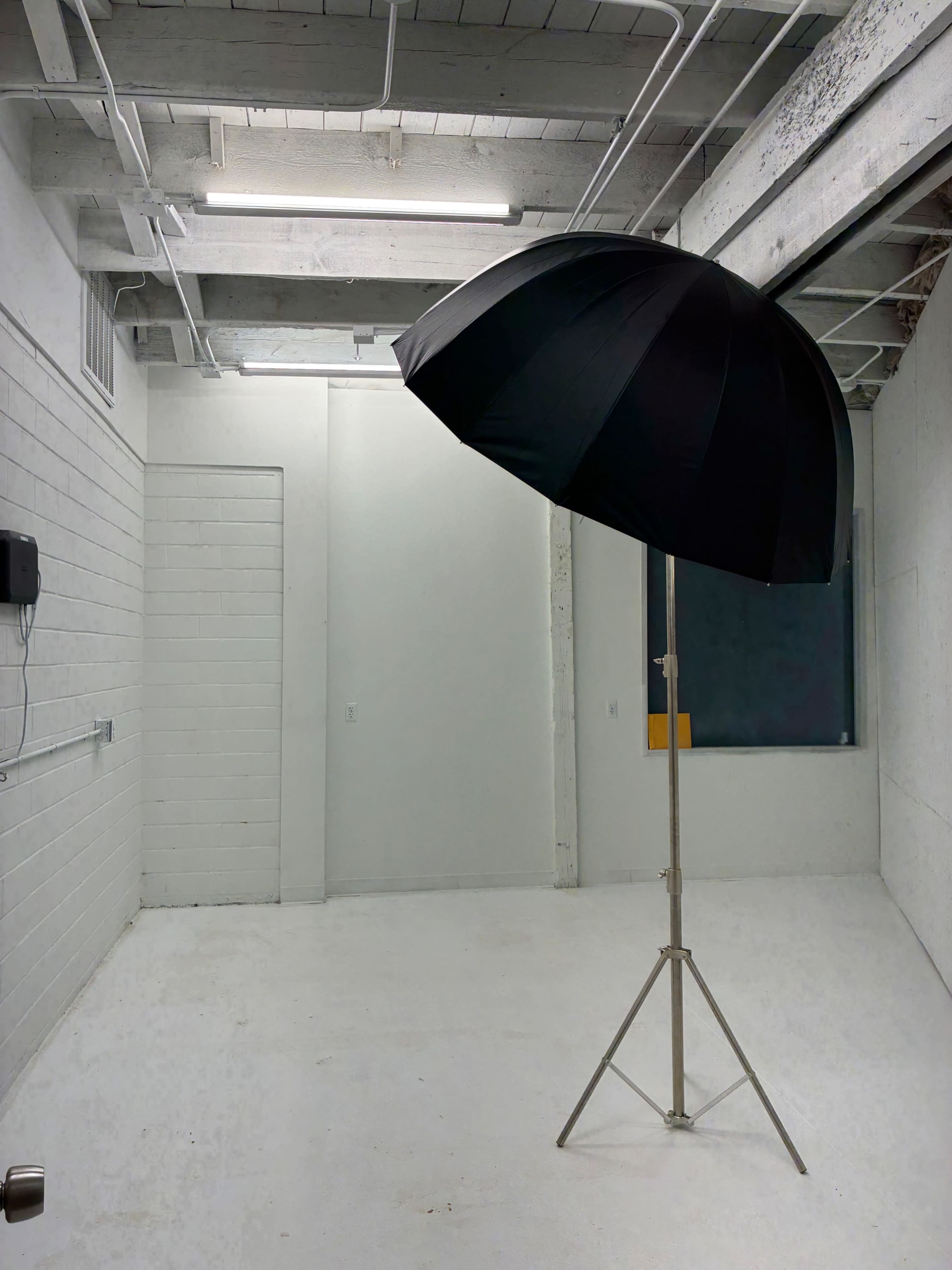 The image shows a large black umbrella on a standing metal frame positioned in a sparse white room with exposed ceiling beams and minimal furnishings.