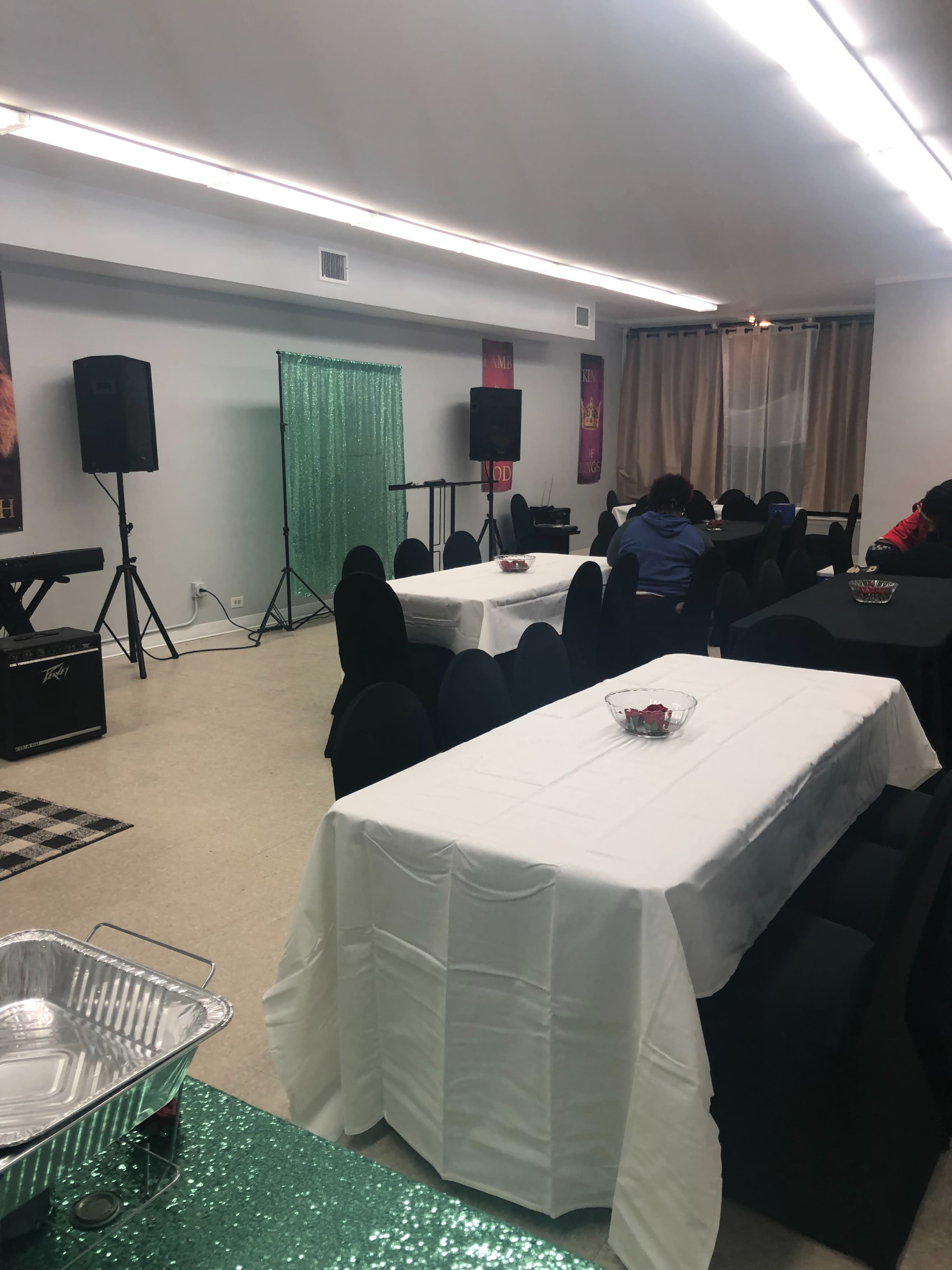 The image shows a spacious event room with tables covered in white tablecloths, green chairs, and a stage area featuring sound equipment and decorative elements.