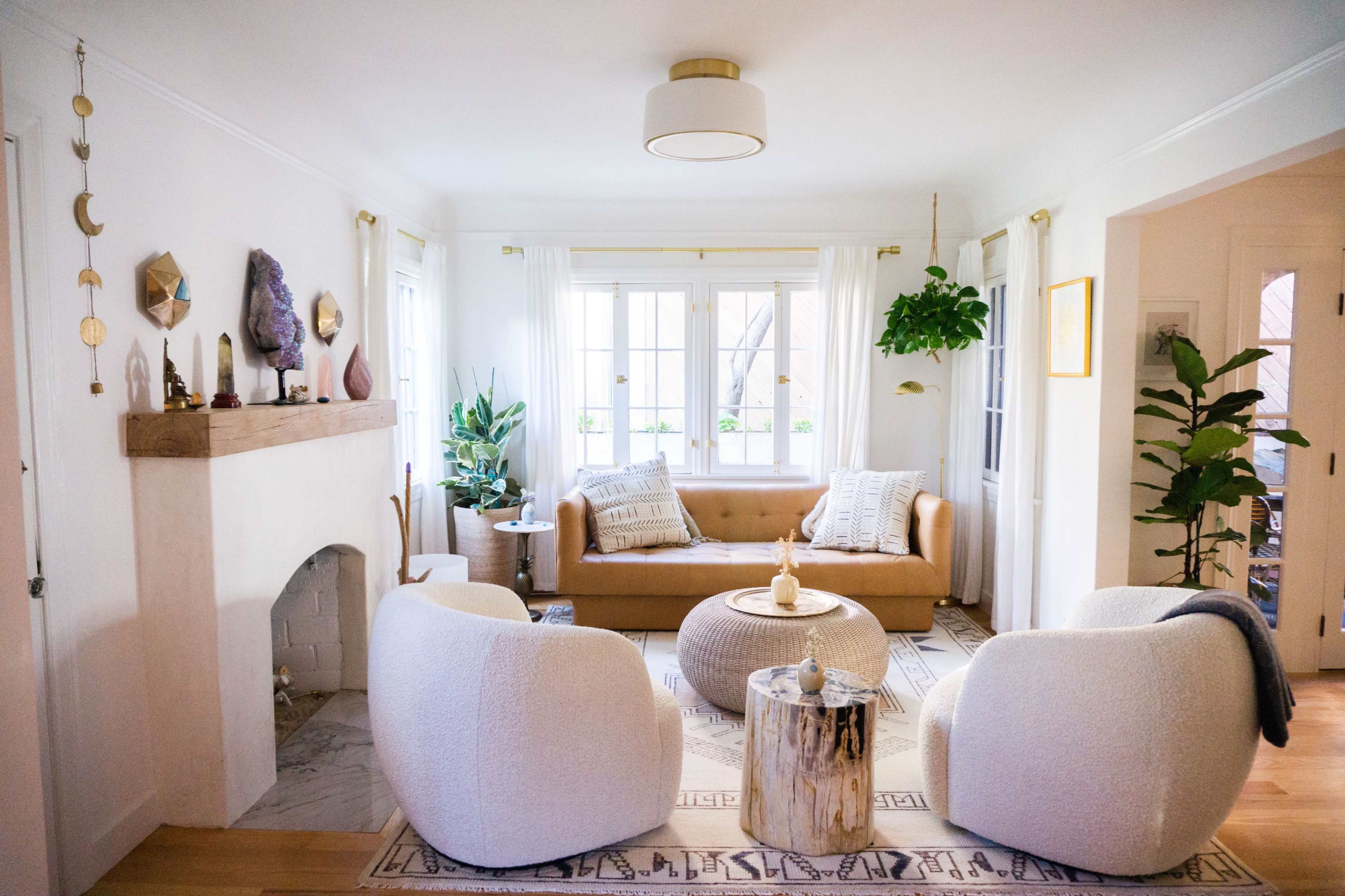 The living room features a tan sofa, two white armchairs, a wooden coffee table, and a fireplace with decorative elements, set against large windows that let in natural light.