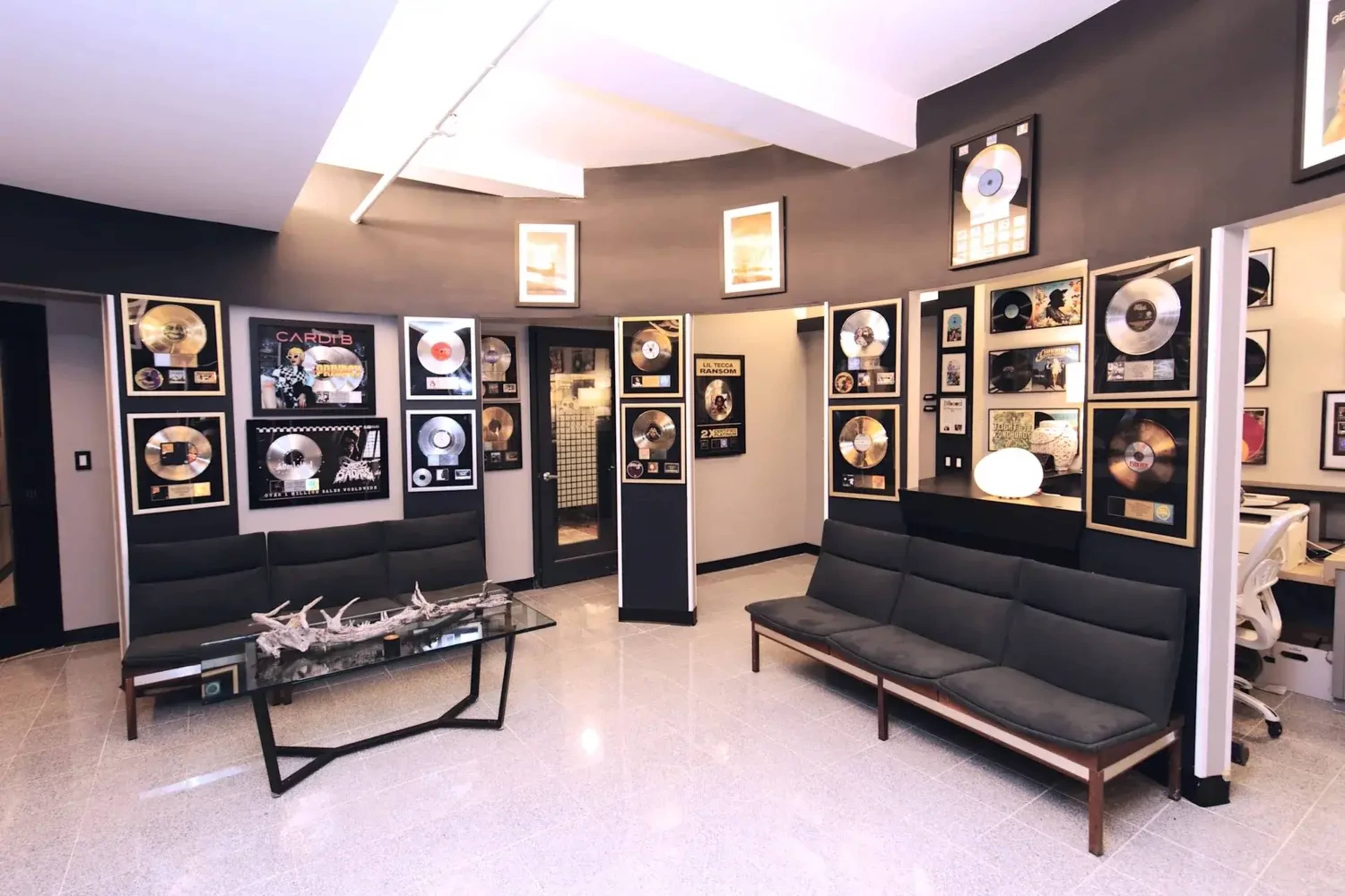 The image shows a modern lounge area adorned with framed gold and platinum music records on the walls, accompanied by sleek black furniture and a glass table.