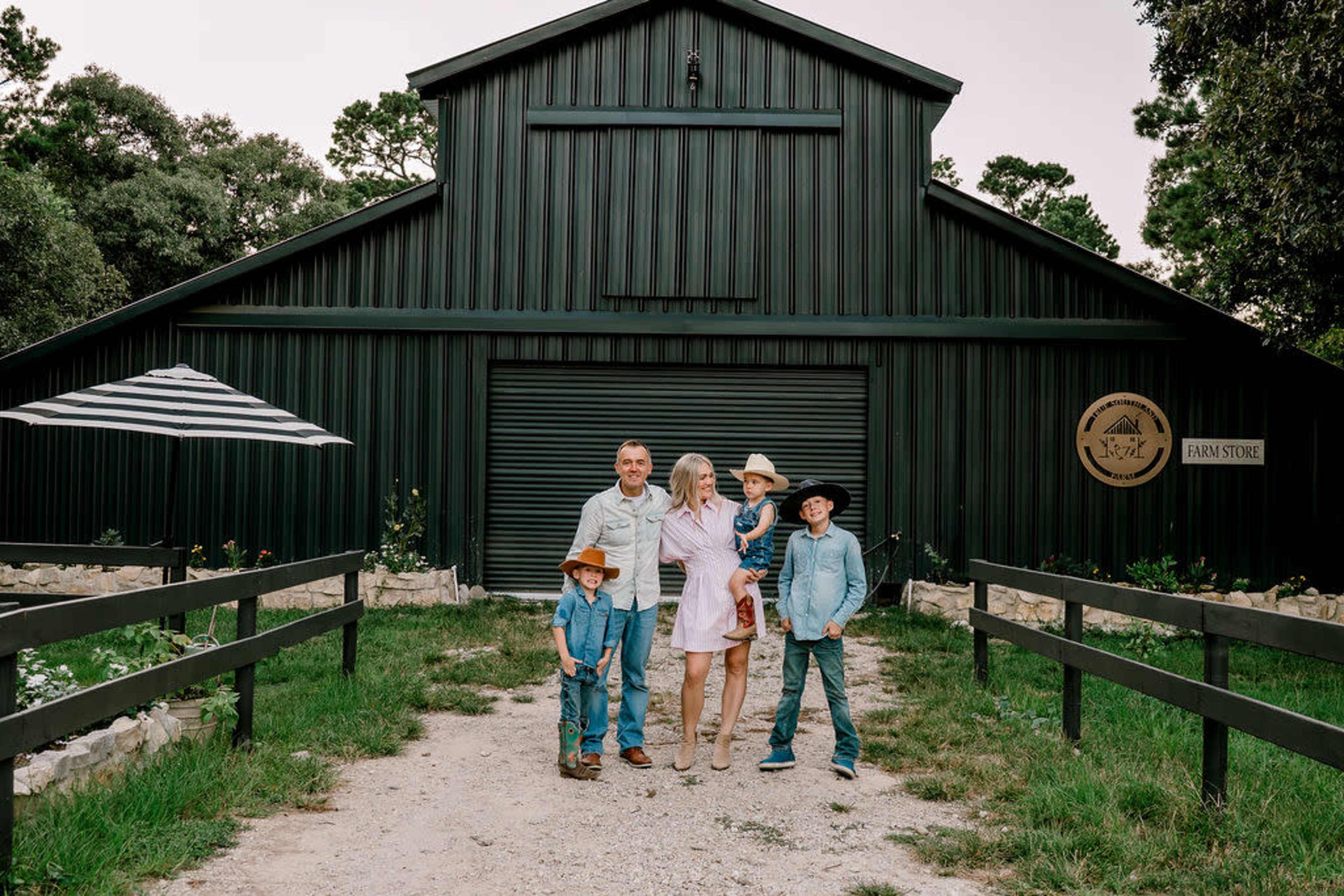 A family of five poses together in front of a large green barn with a sign reading "Farm Store."