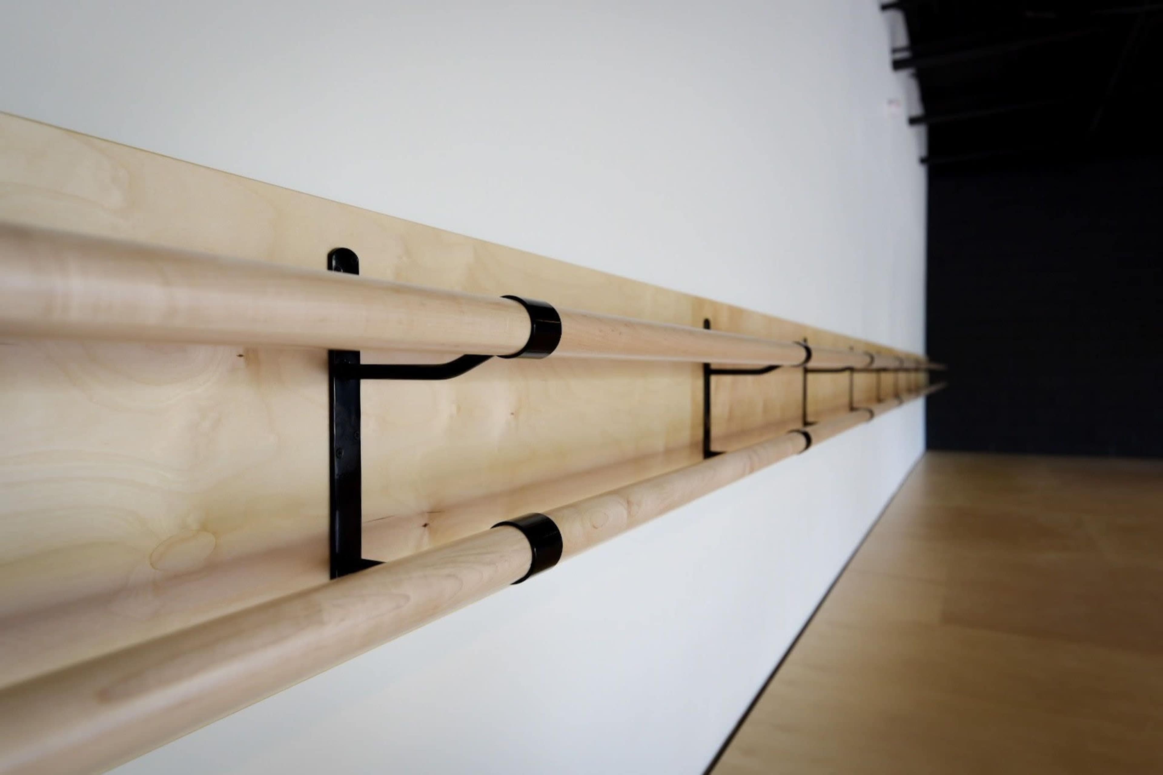 The image shows a line of wooden ballet barres mounted on a wall in a studio with a hardwood floor.