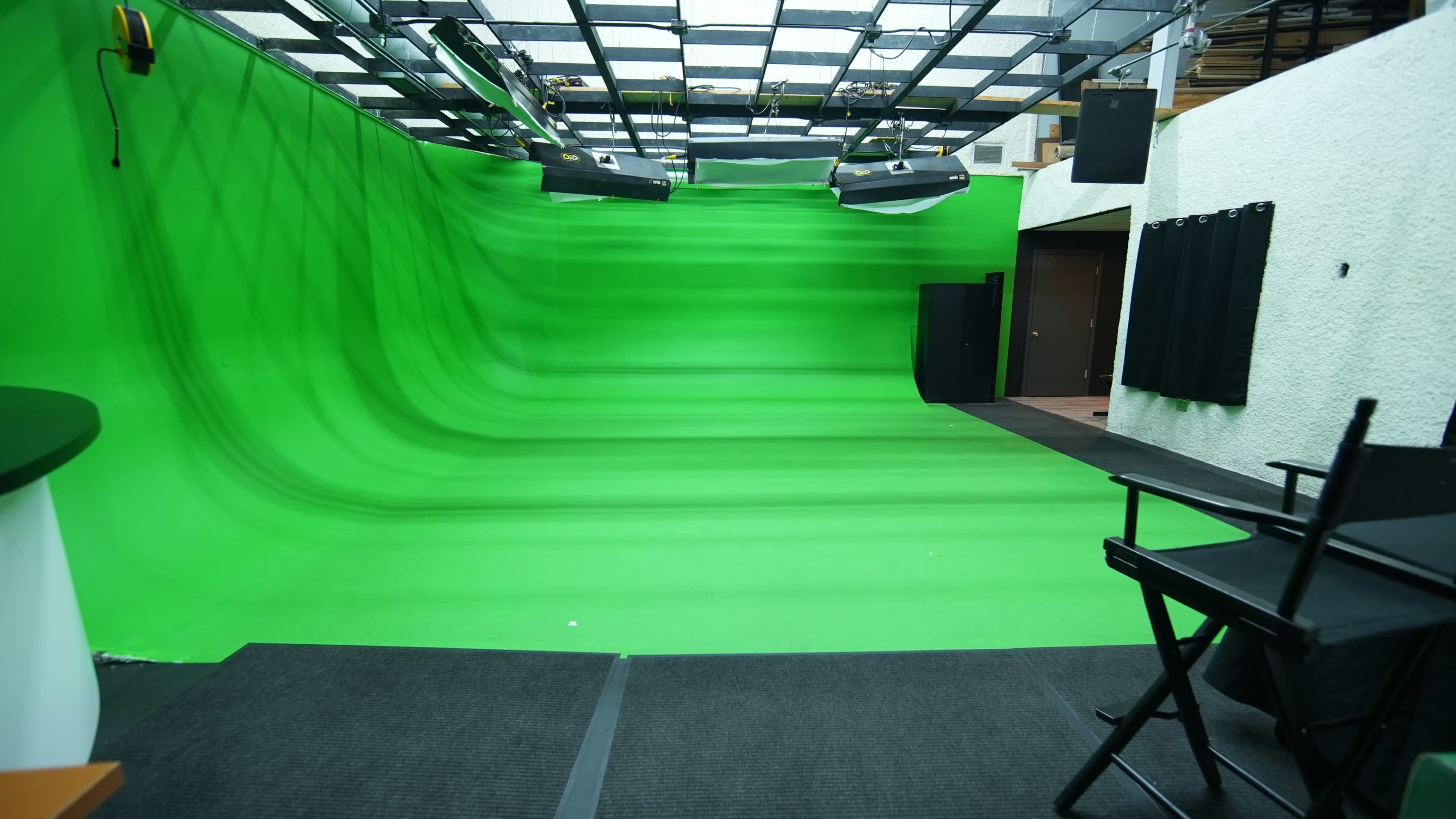The image shows a green screen studio with a curved green backdrop, lighting fixtures overhead, and a director's chair positioned on a black mat.