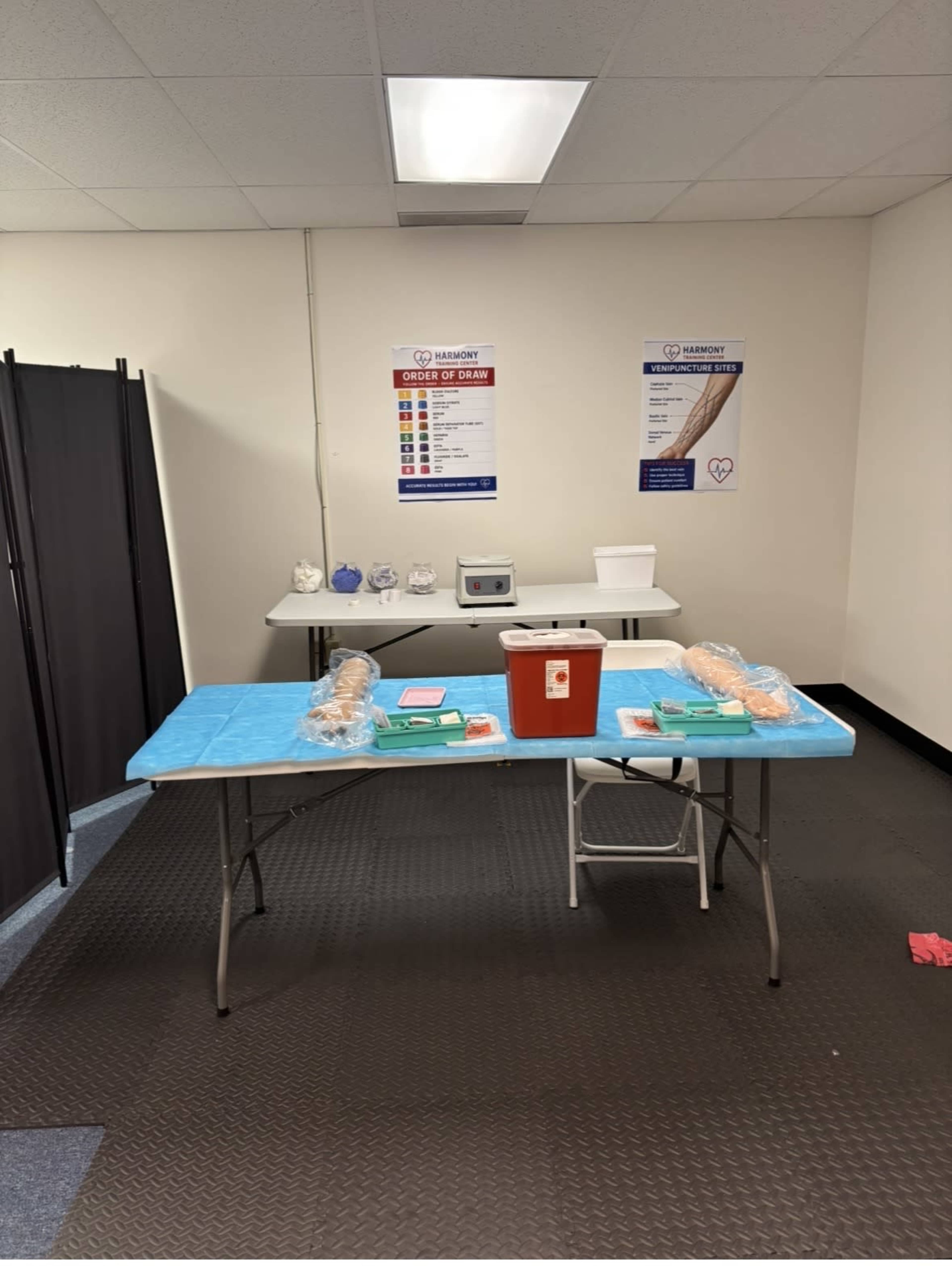 The image shows a clean, organized medical room featuring a blue table with medical supplies, a red biohazard container, and additional equipment in the background.