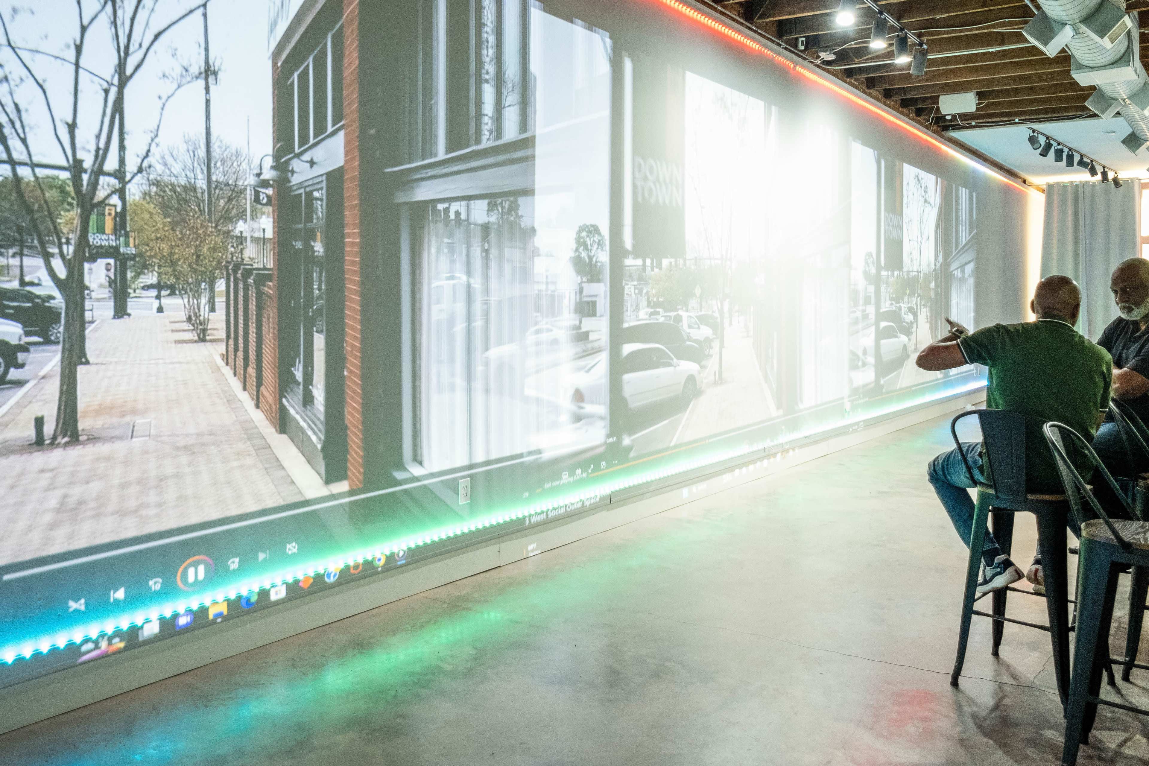 A long wall displays a projected cityscape while two people sit at a bar in the foreground.
