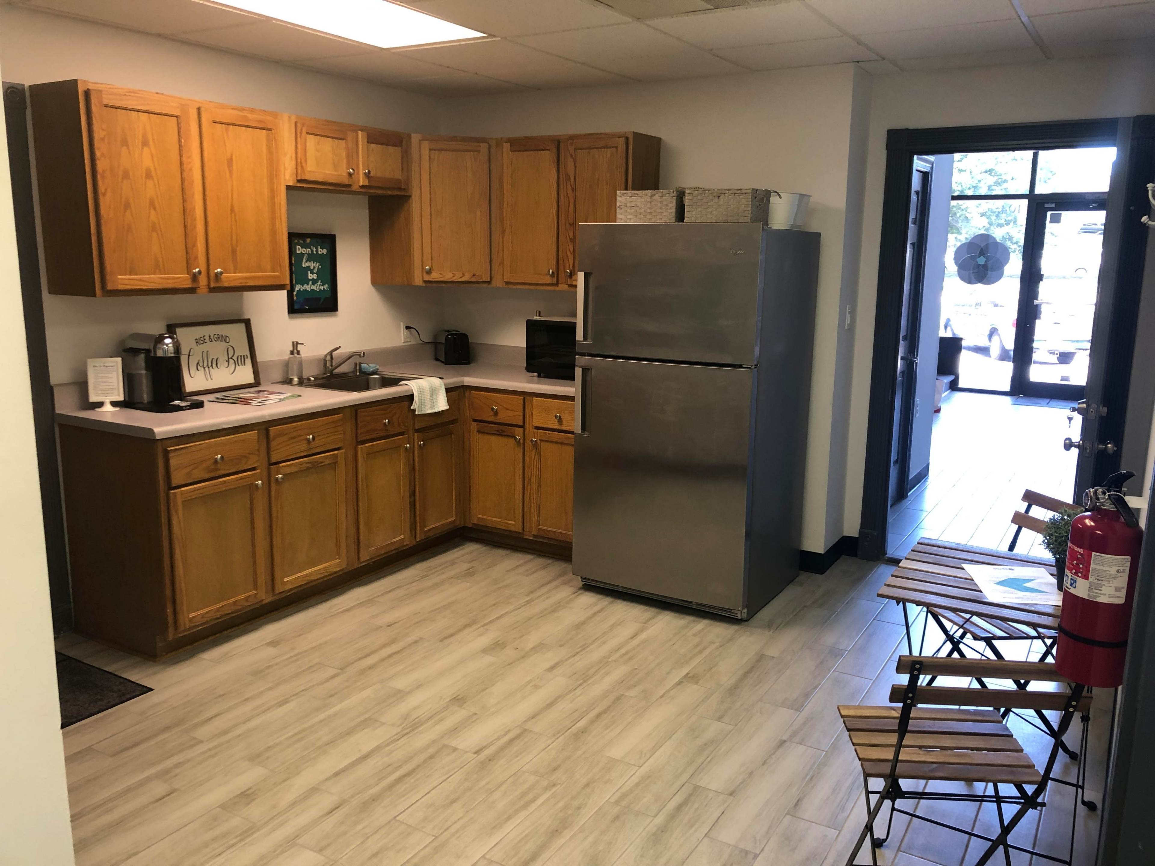 A kitchen with wooden cabinets, a stainless steel refrigerator, a small table with chairs, and a doorway leading outside.