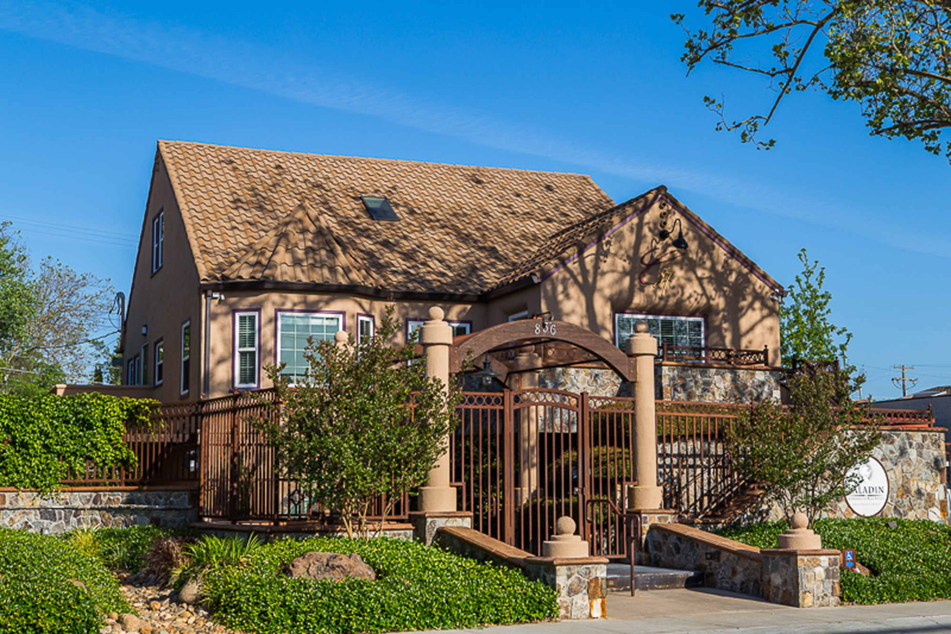 A two-story house with a tan exterior and a brown roof is surrounded by a stone wall and iron gate, set against a clear blue sky.