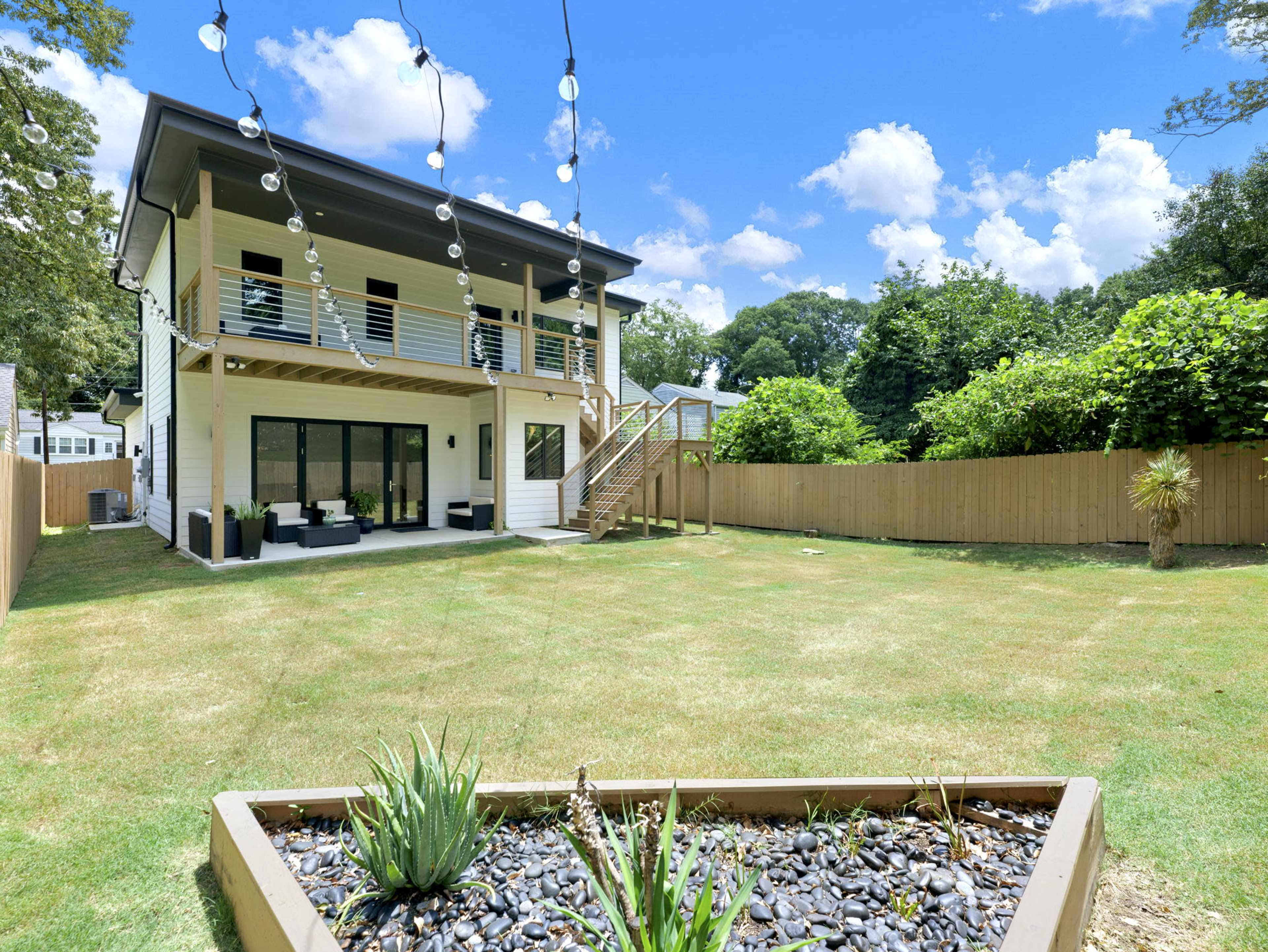 The image shows a two-story house with a spacious backyard featuring a patio, a landscaped area with rocks and plants, and string lights overhead.
