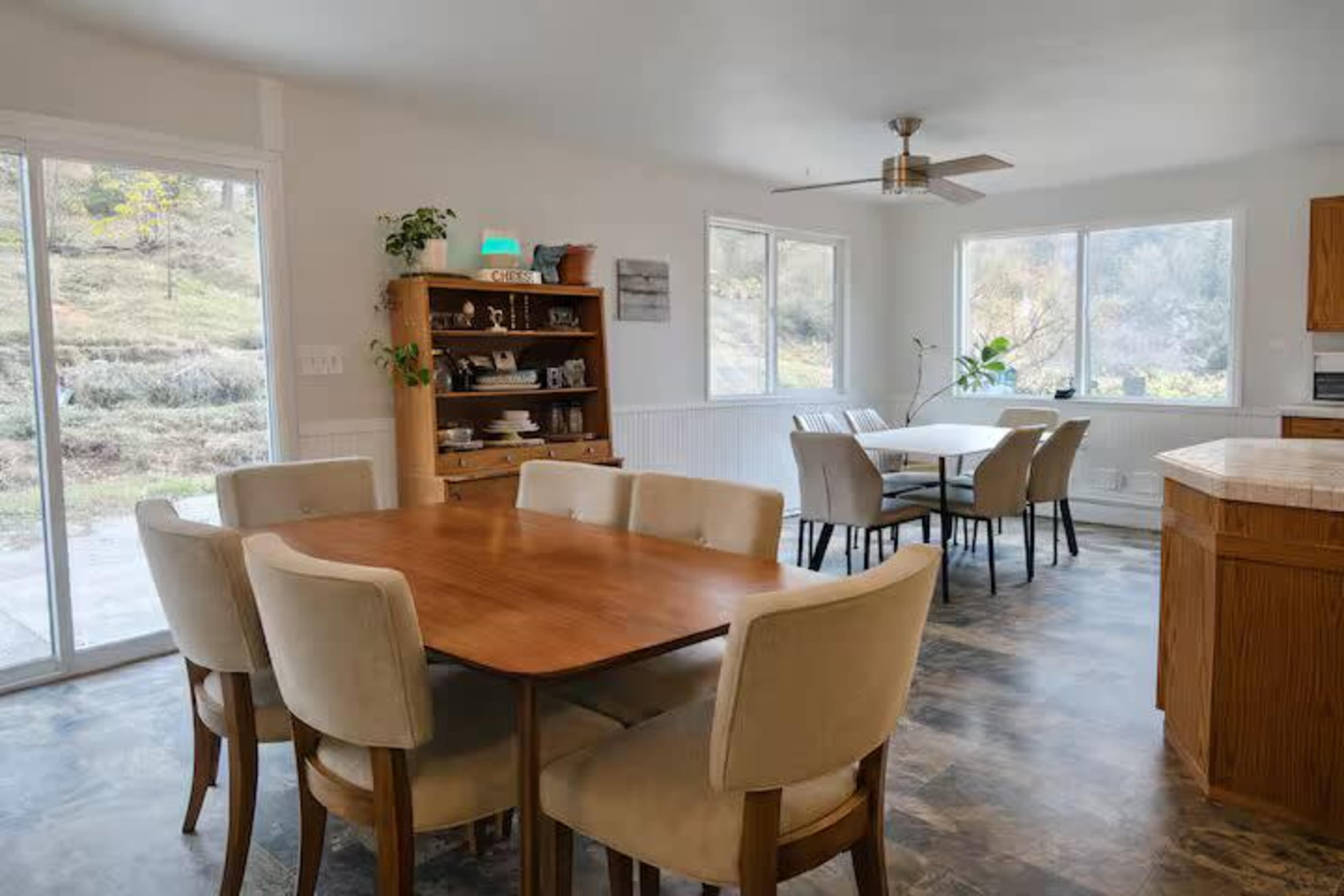 The image shows a modern dining area featuring a wooden table with upholstered chairs, a large window letting in natural light, and a shelf filled with decorative items.