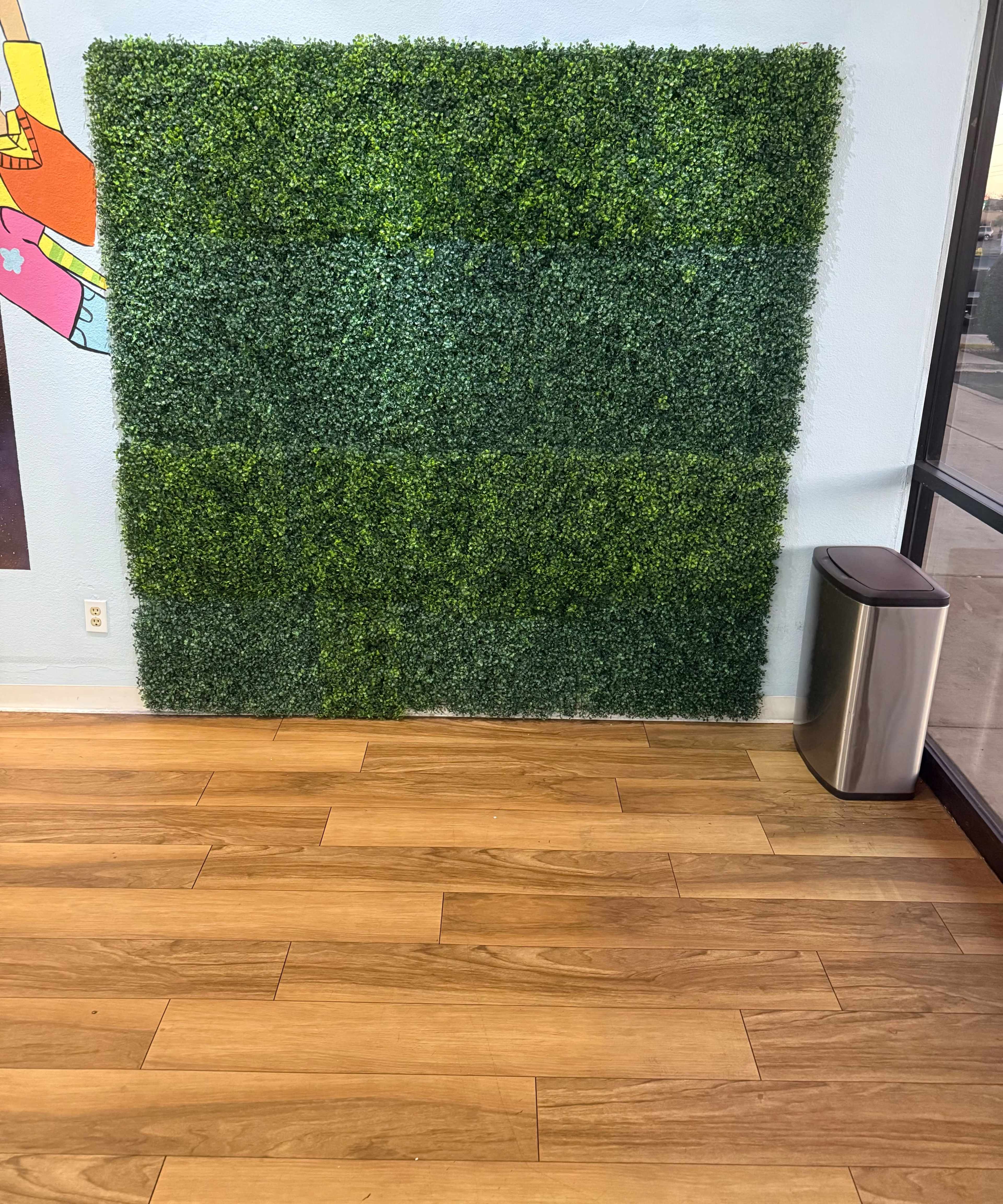 The image shows a green artificial grass wall next to a silver trash can on a wooden floor inside a room.