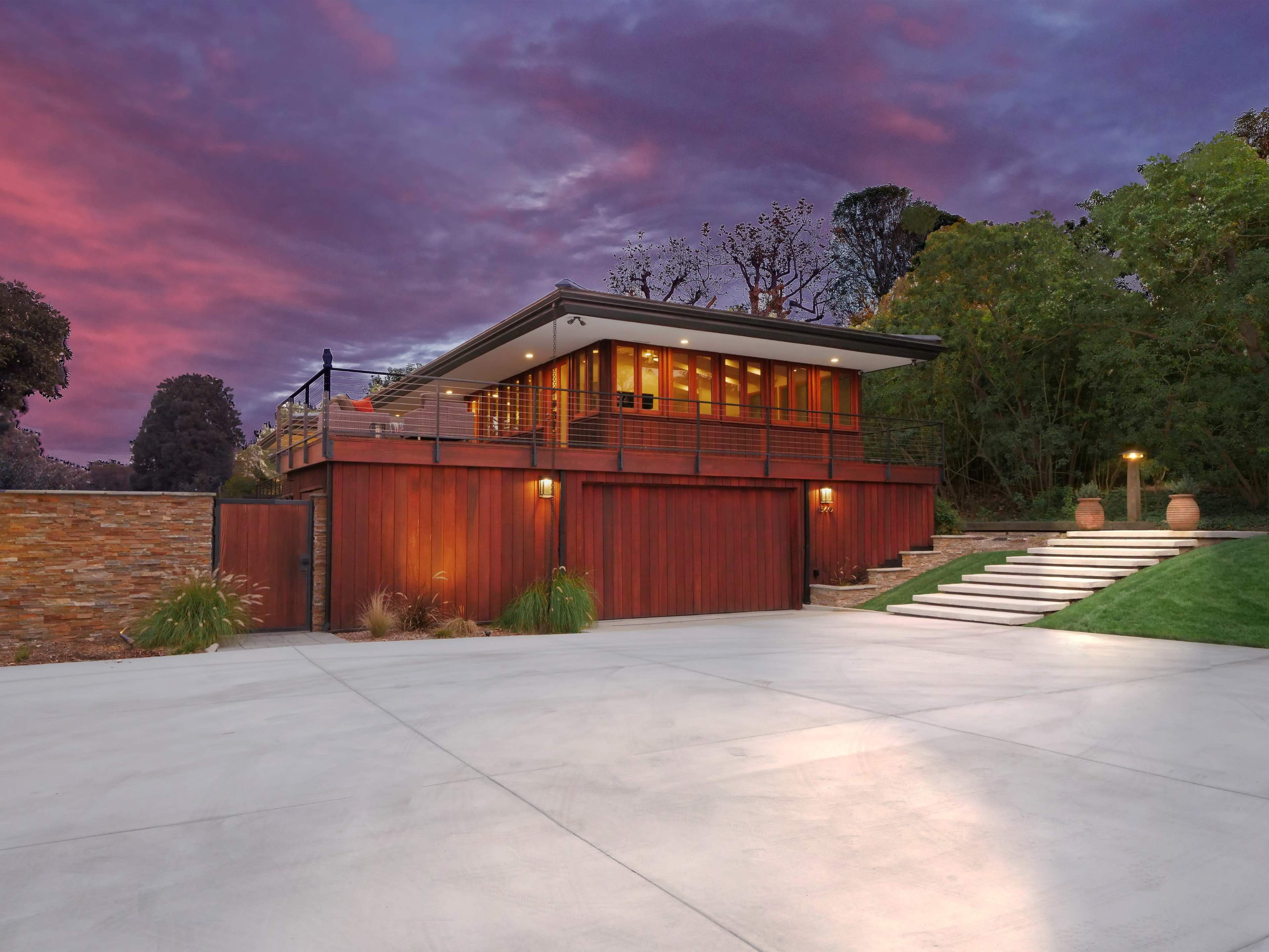 A modern wooden house with large windows sits atop a landscaped driveway, illuminated by evening light against a colorful sky.