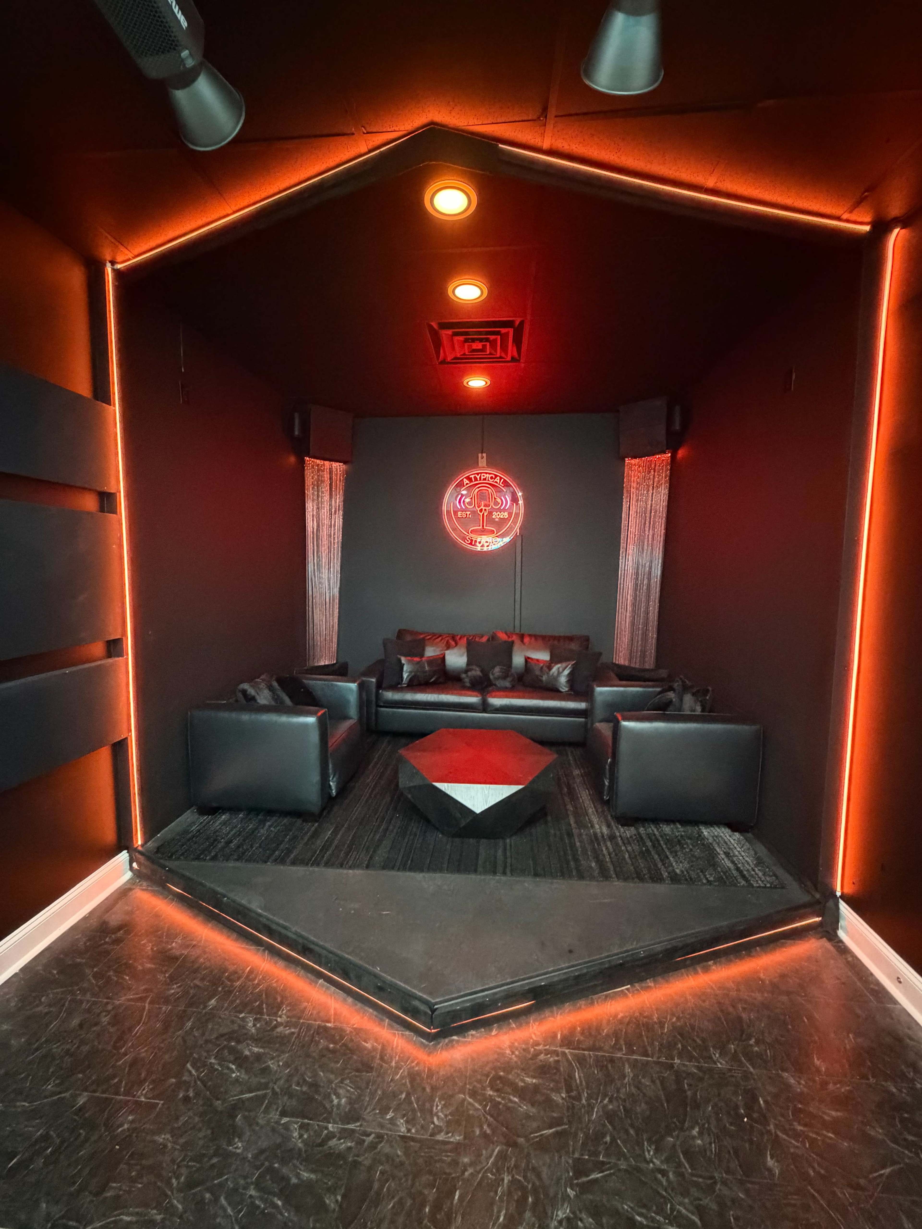 The image shows a modern lounge area with black walls, red accent lighting, and sleek black furniture arranged around a geometric coffee table.