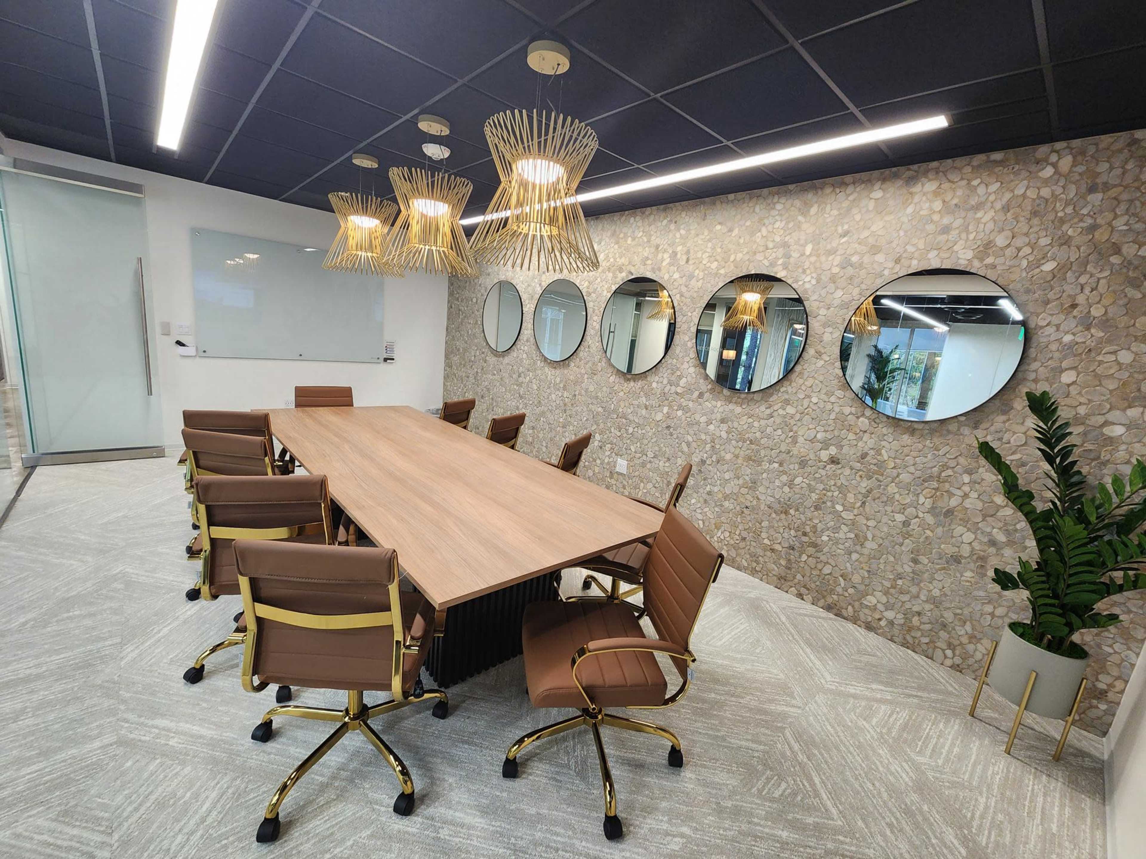 A conference room features a long wooden table surrounded by brown chairs, with a stone wall displaying circular mirrors and modern light fixtures above.